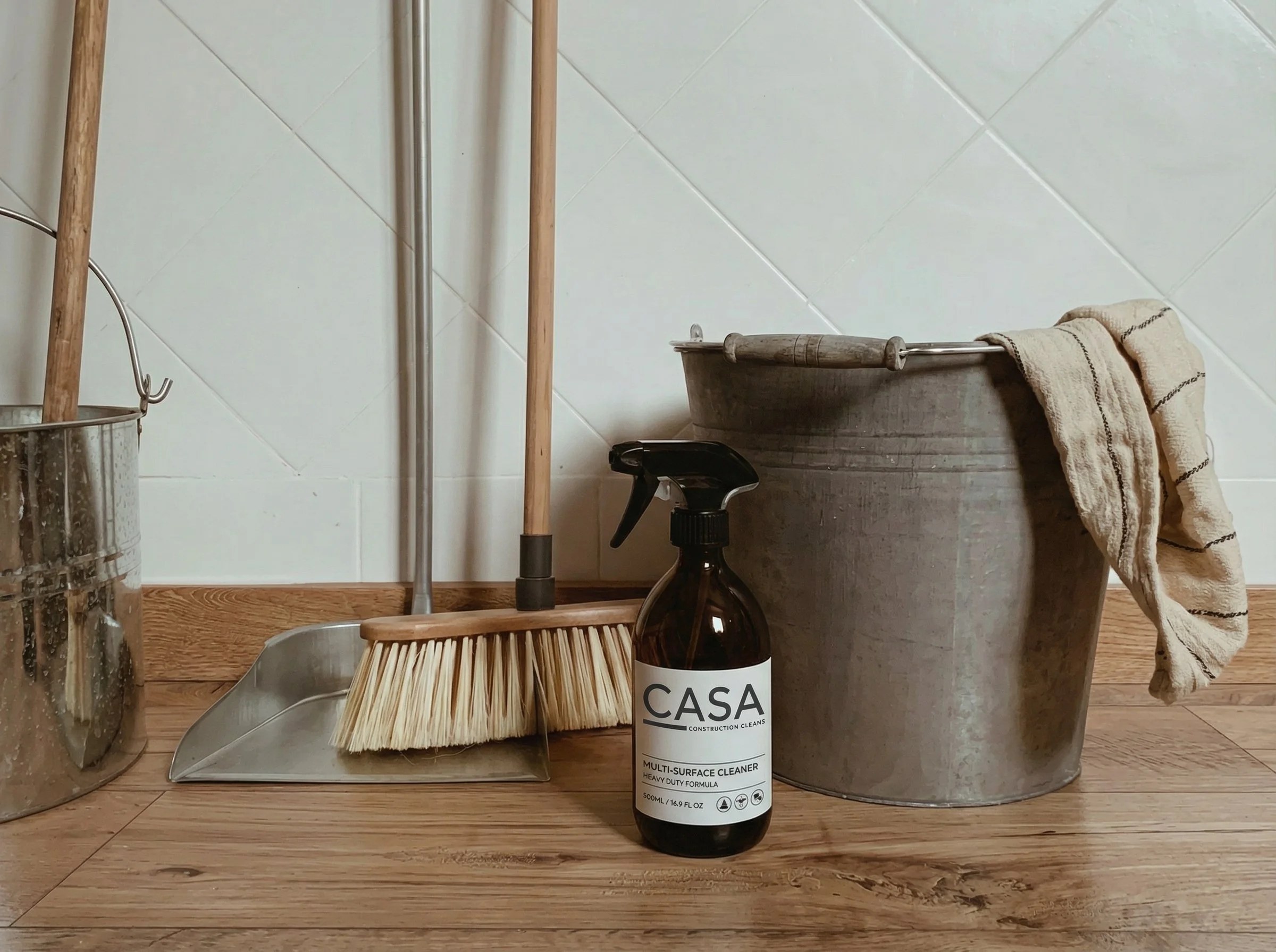 A collection of cleaning tools and supplies on a wooden floor, including a metal bucket, a straw brush, a spray bottle labeled 'Casa Construction Cleans', a large metal container with a towel draped over it, a sprayer, a mop, and a broom against a white tiled wall.