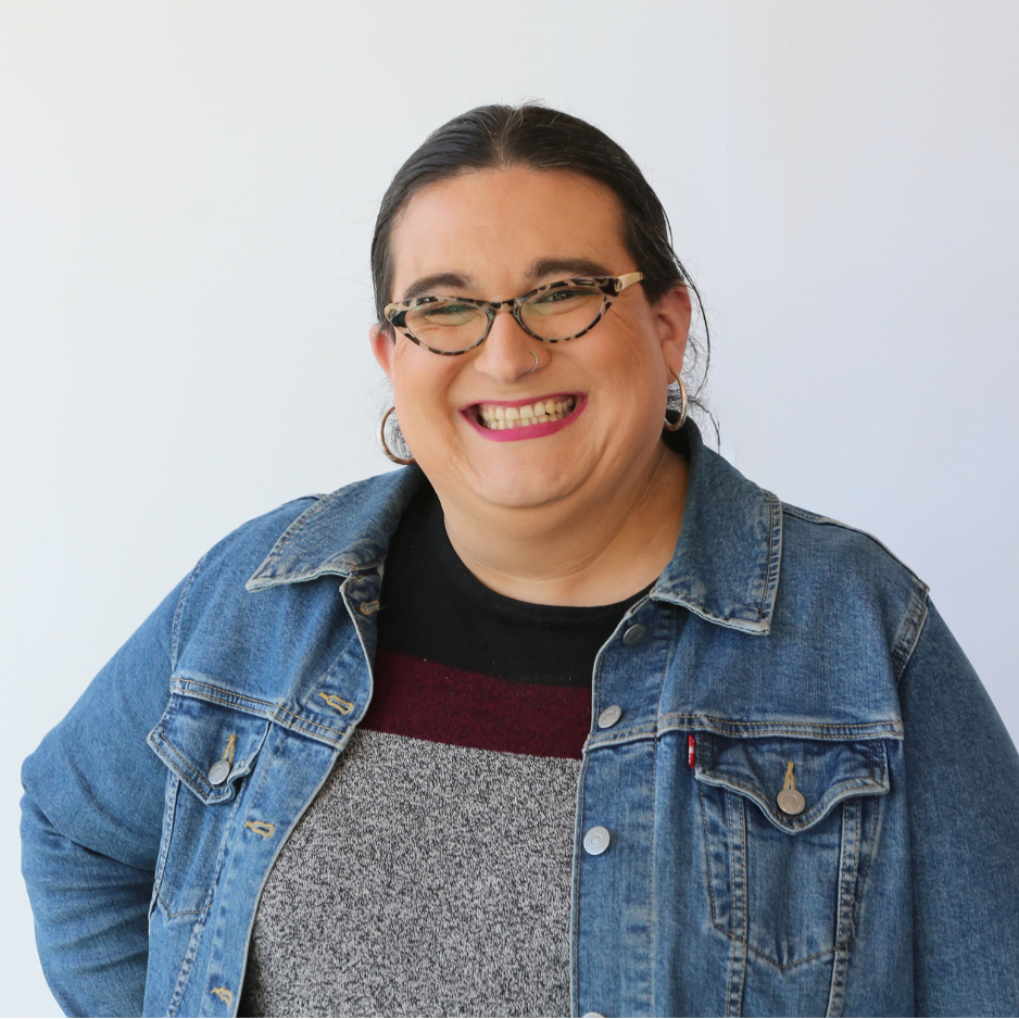 A woman with dark hair, wearing glasses, a denim jacket, and a striped shirt, smiling and posing against a white background.