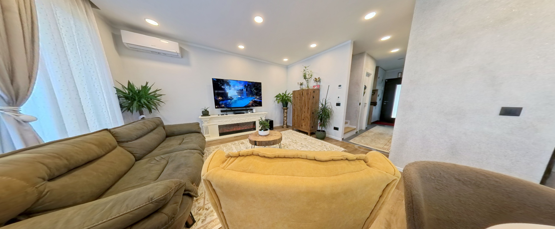 Living room with a beige sofa, a yellow armchair, a white fireplace mantle with a flat-screen TV, indoor plants, wooden furniture, and a patterned rug.