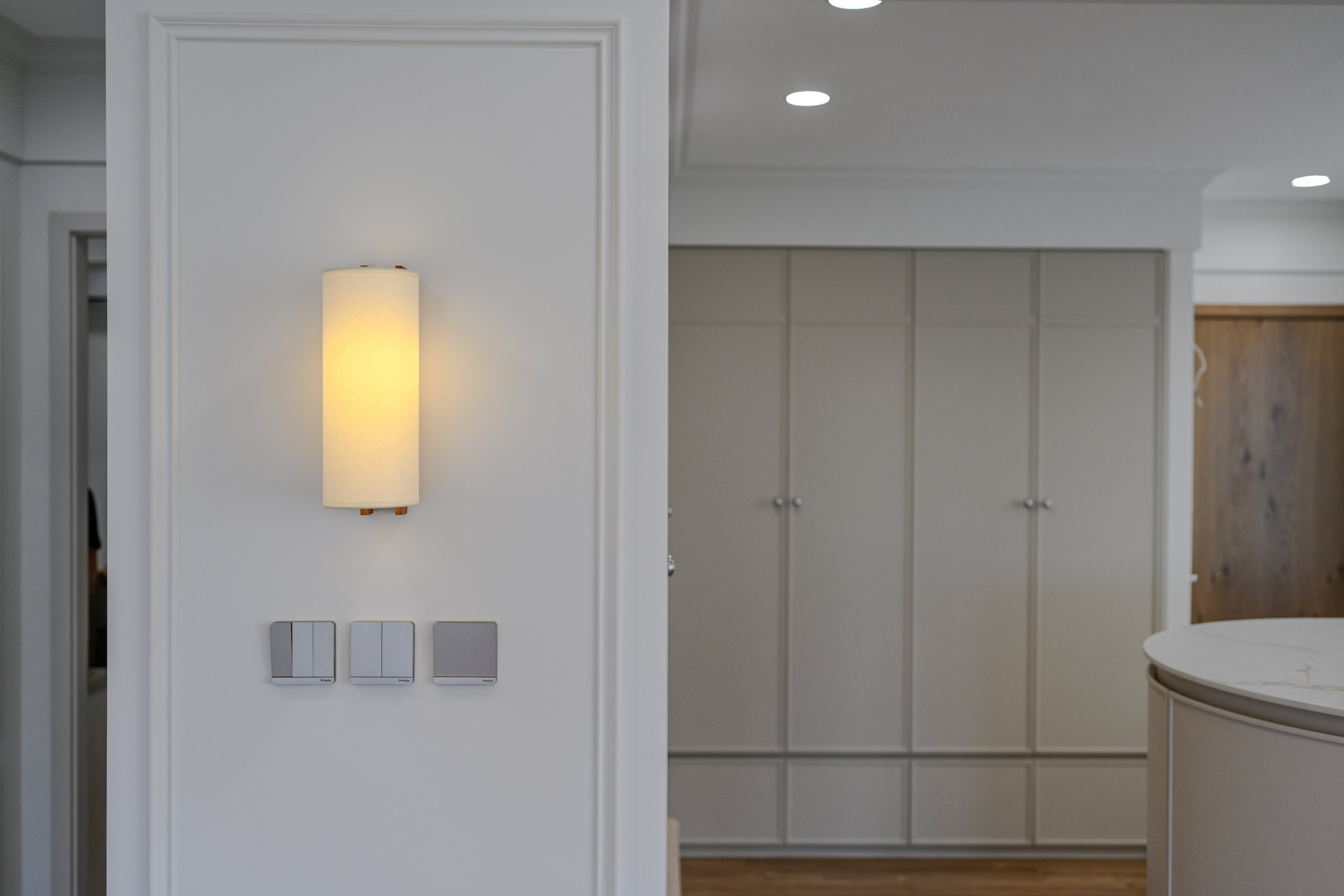 Interior of a modern home with a wall-mounted lamp, three light switches, and a view of a corridor with built-in cabinets and a wooden door.