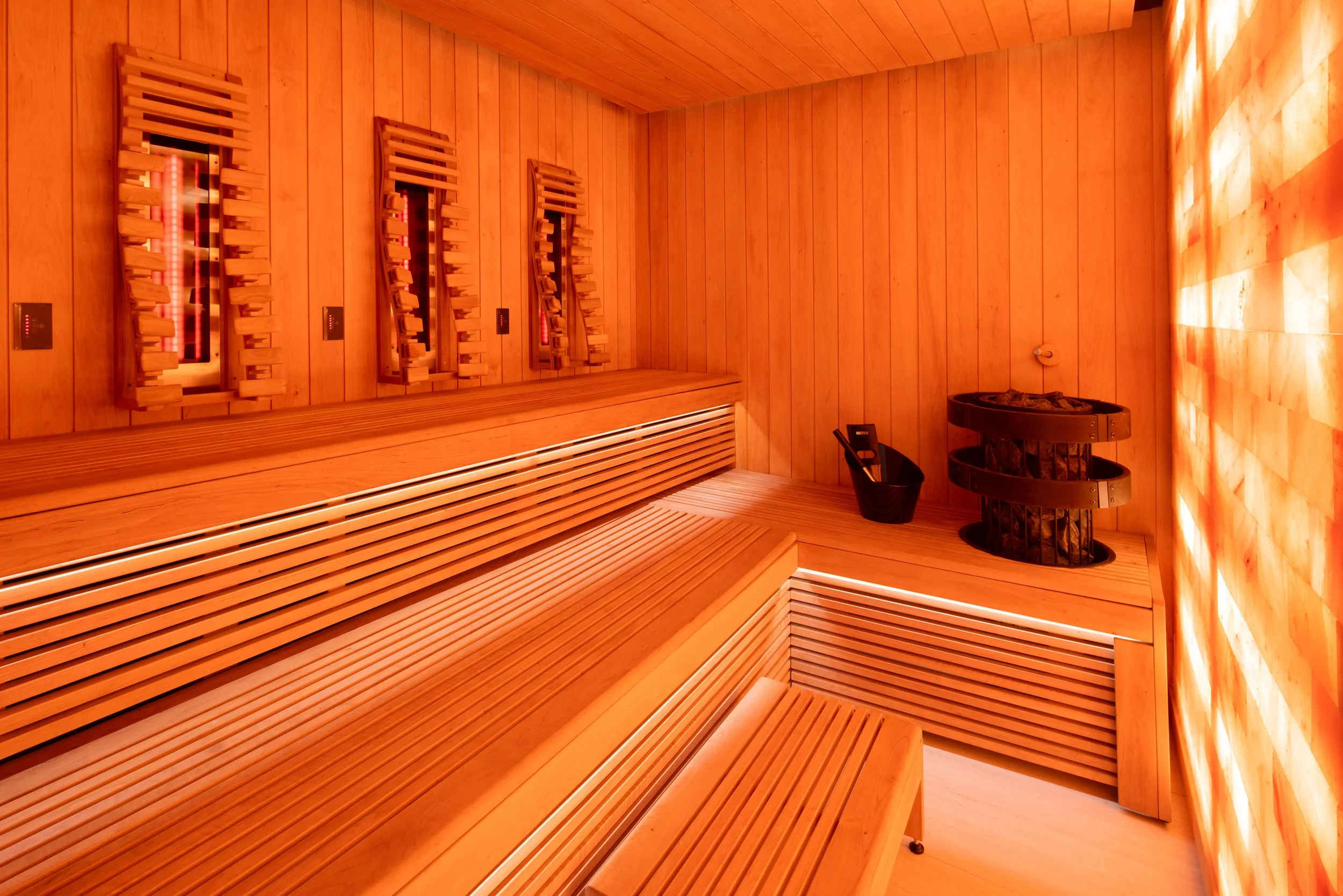 A wooden sauna room with benches, sauna controls, a bucket, and a heater with stones inside.