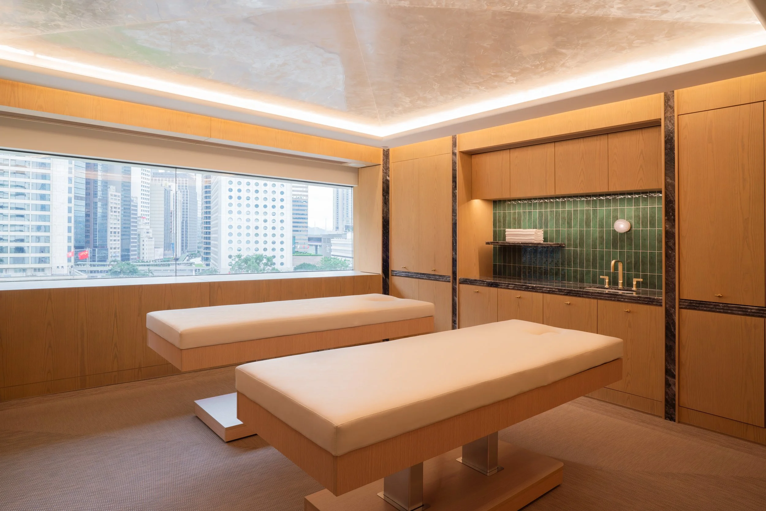 A modern spa or massage room with two massage tables and a large window showing a cityscape with high-rise buildings.