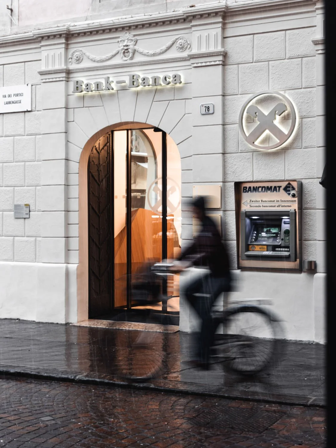 Ein Gebäude mit einem Eingang, der als Bank gekennzeichnet ist, darunter ein Geldautomat. Es ist eine Person zu sehen, die auf einem Fahrrad vorbeifährt.