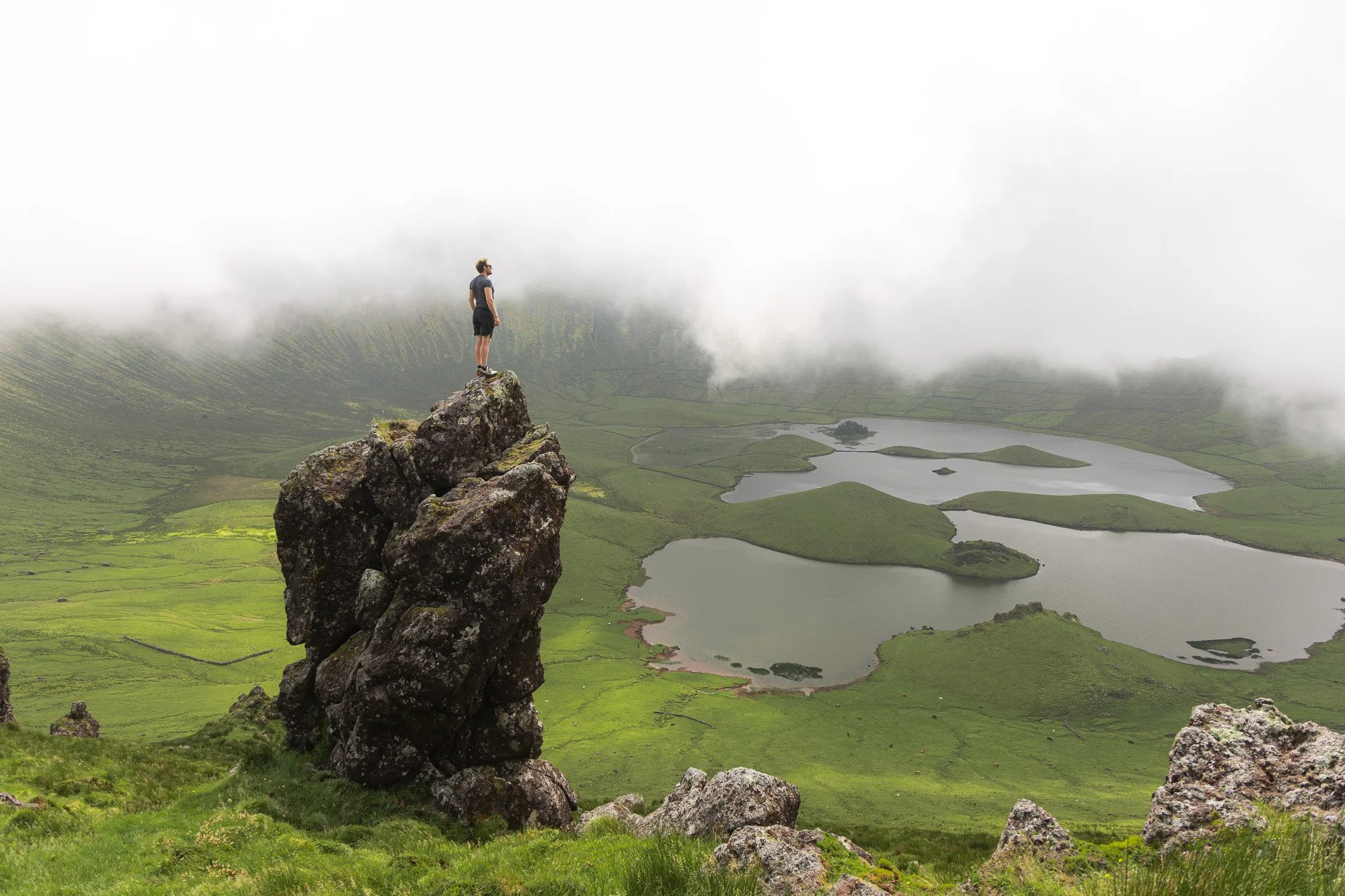 Eine Person steht auf einem großen Felsen in einer grünen, bergigen Landschaft mit Seen, umgeben von Wolken.