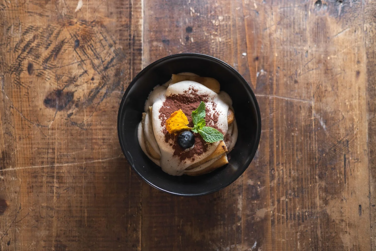 Eine kleine Schüssel mit Tiramisú, garniert mit Schokoladenpulver, Minzblättern, einer Heidelbeere und einem kleinen Stück Orange, auf einem holzernen Tisch.