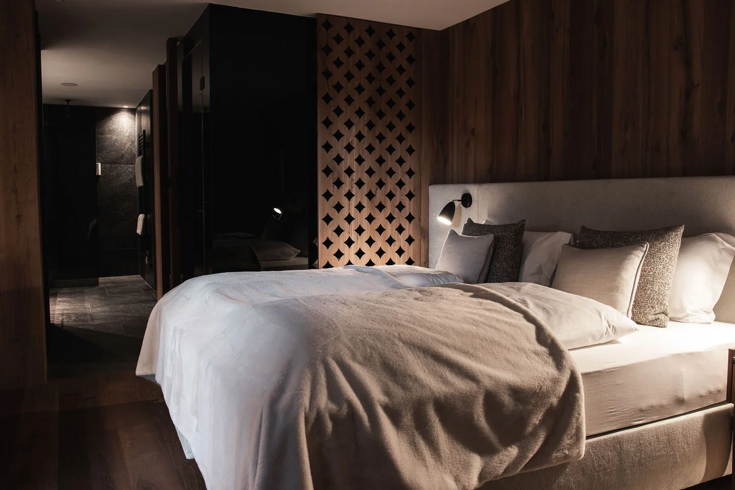 Ein modernes Schlafzimmer mit einem großen, gemütlichen Bett, mehrere Kissen, einem Nachttisch mit einer Leselampe und dunklem Holzpaneel im Hintergrund.