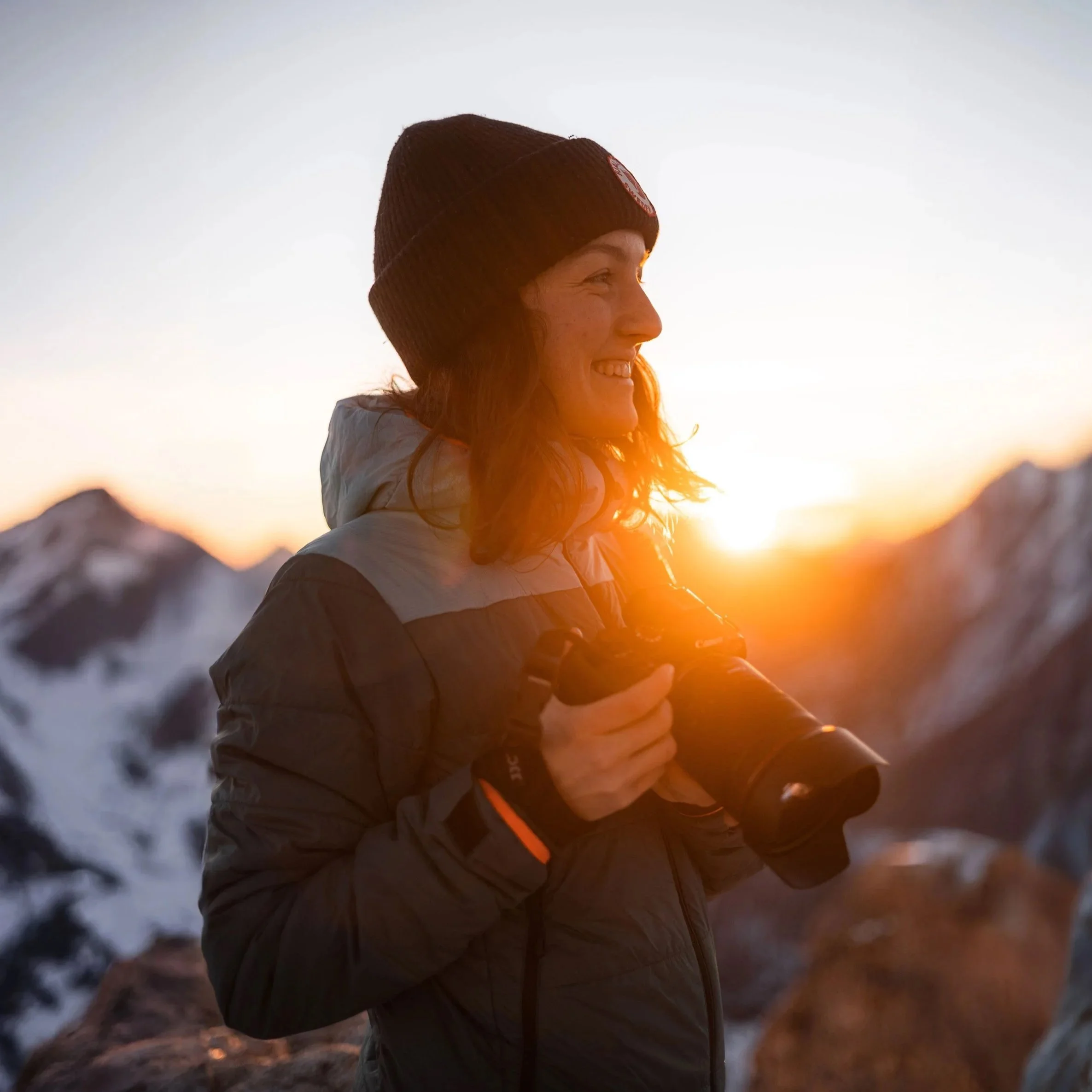 Frau mit Kamera beim Sonnenaufgang in den Bergen, trägt Winterjacke und Mütze.