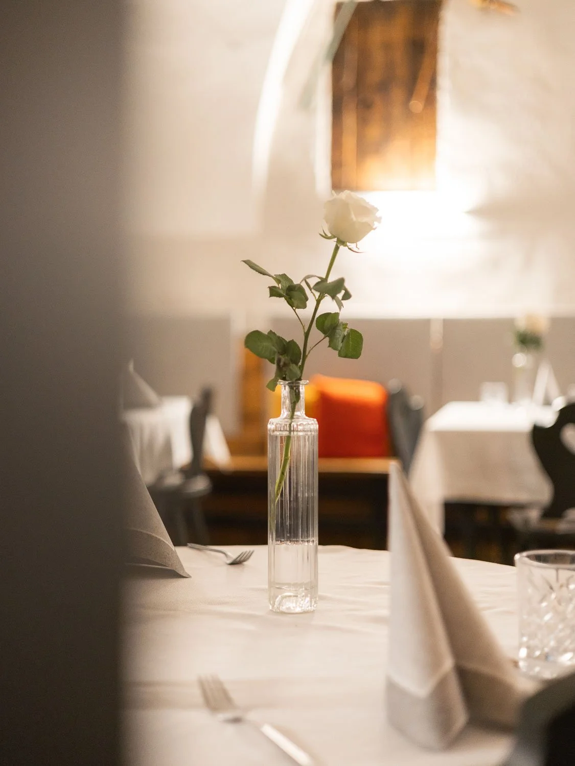 Eine weiße Rose in einer klaren Vase auf einem Esstisch in einem Restaurant, im Hintergrund Fenster und aufgestellte Tische mit Besteck und Gläser.