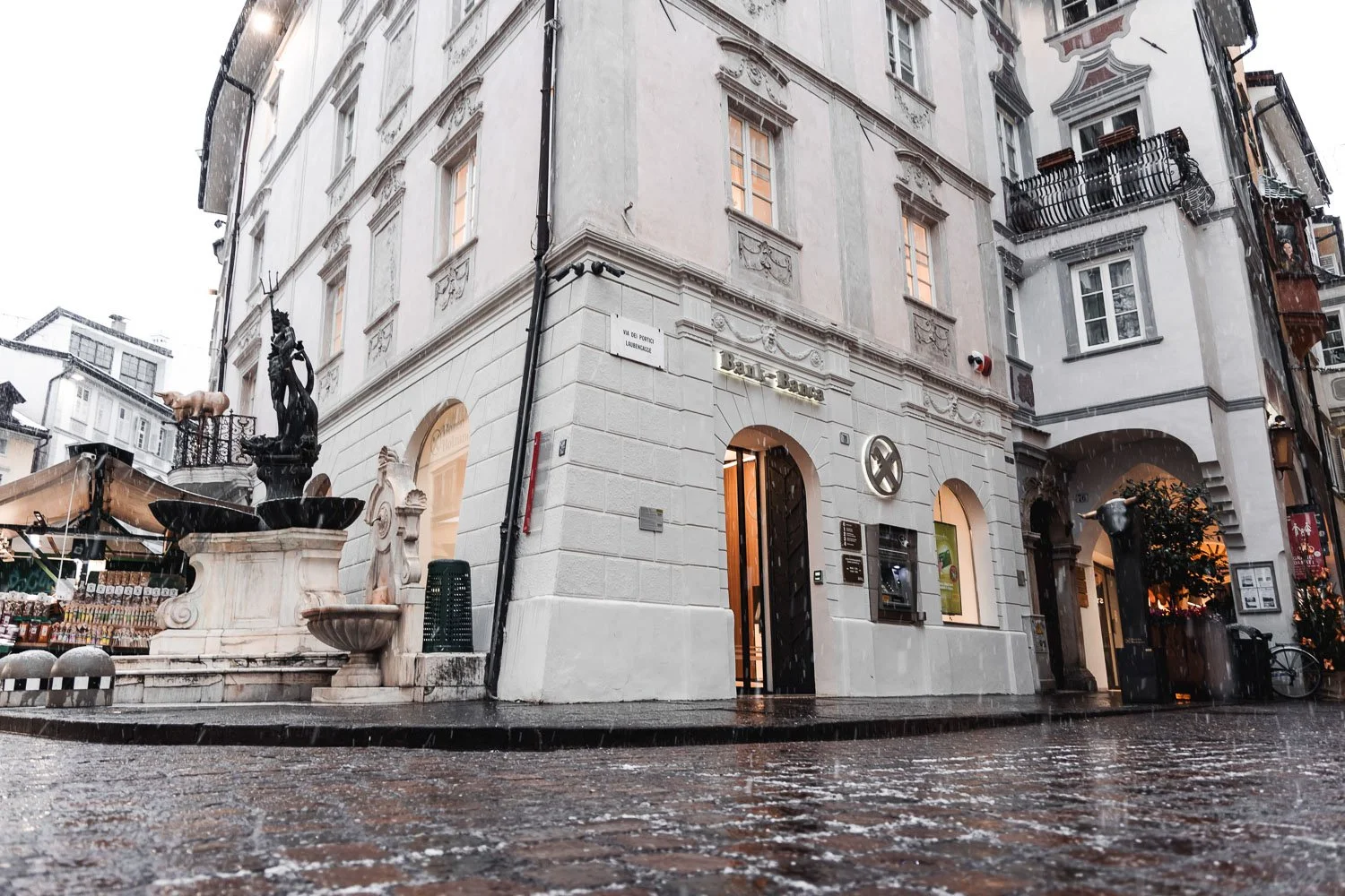 Straßenansicht eines alten, weißen Gebäudes mit mehreren Fenstern und Balkonen, einem Brunnen mit Skulptur vor dem Gebäude, bei regnerischem Wetter.