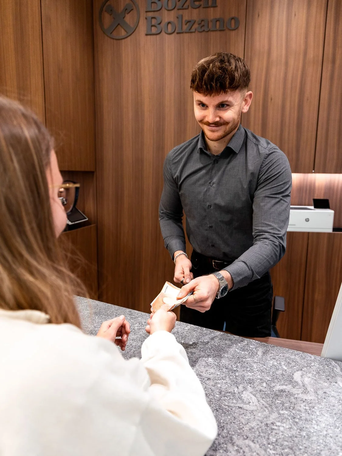 Ein Mann am Empfang gibt einer Frau Geld aus, im Hintergrund ein Holz-Wand und ein Schild mit der Aufschrift "Bozen".