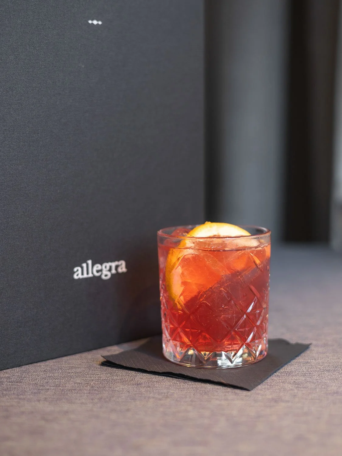 Ein Glas mit rotem Cocktail und Zitrone, auf einer schwarzen Serviette auf einem grauen Tischtuch, im Hintergrund eine schwarze Speisekarte mit der Aufschrift "allegra".