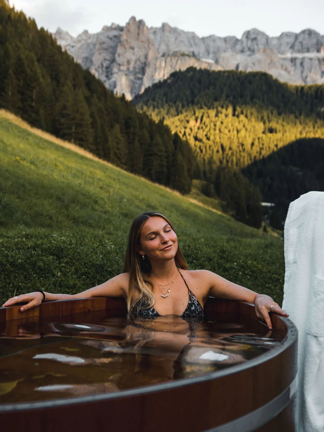 Eine junge Frau entspannt in einer Holzzuber-Badewanne im Freien mit Blick auf grüne Hügel, Wälder und Berggipfel im Hintergrund bei Sonnenuntergang.