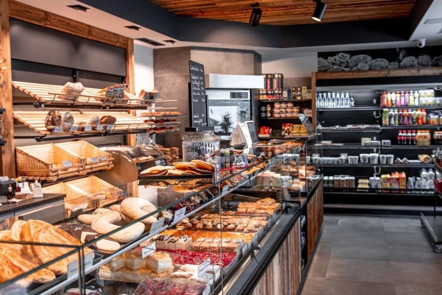 Bäckerei mit ausgestellten Brötchen, Kuchen und Backwaren aus Holzregalen und einem Glasvitrine, sowie Regalen mit Getränken und Lebensmitteln im Hintergrund.