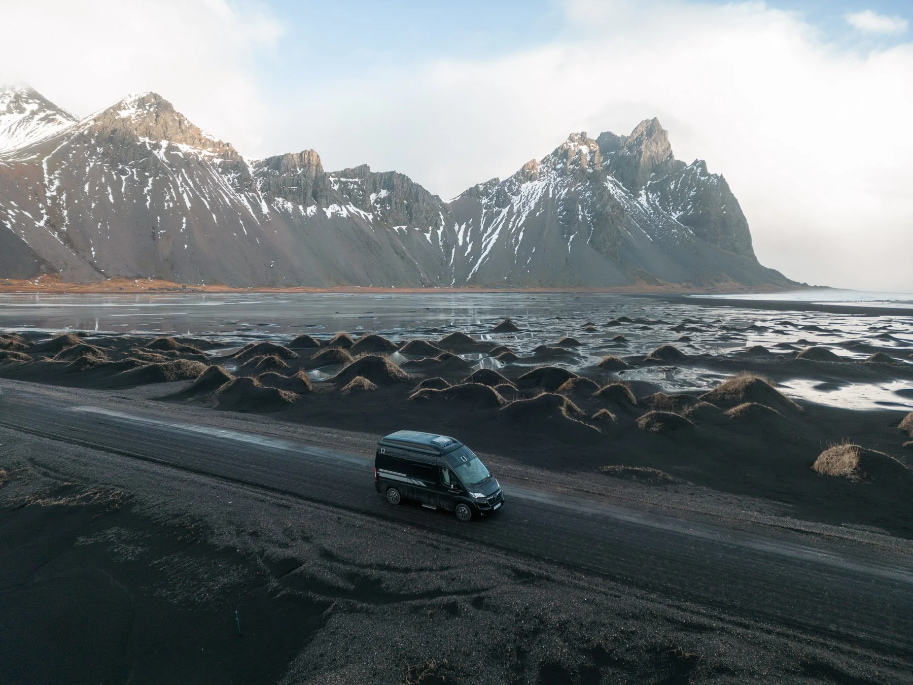 Ein schwarzer Van fährt auf einer Straße entlang einer schwarzen Sandküste mit kleinen Hügeln, mit hohen, schneebedeckten Bergen im Hintergrund unter einem bewölkten Himmel.
