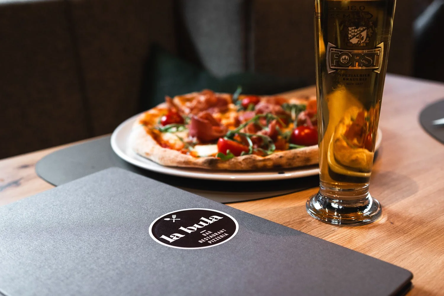Eine Pizza mit Tomaten, Rucola und Schinken steht auf einem Teller auf einem Tisch. Neben der Pizza steht ein Glas Bier. Auf dem Tisch liegt eine speisekartenartige Karte mit dem Schriftzug 'La Bula'.