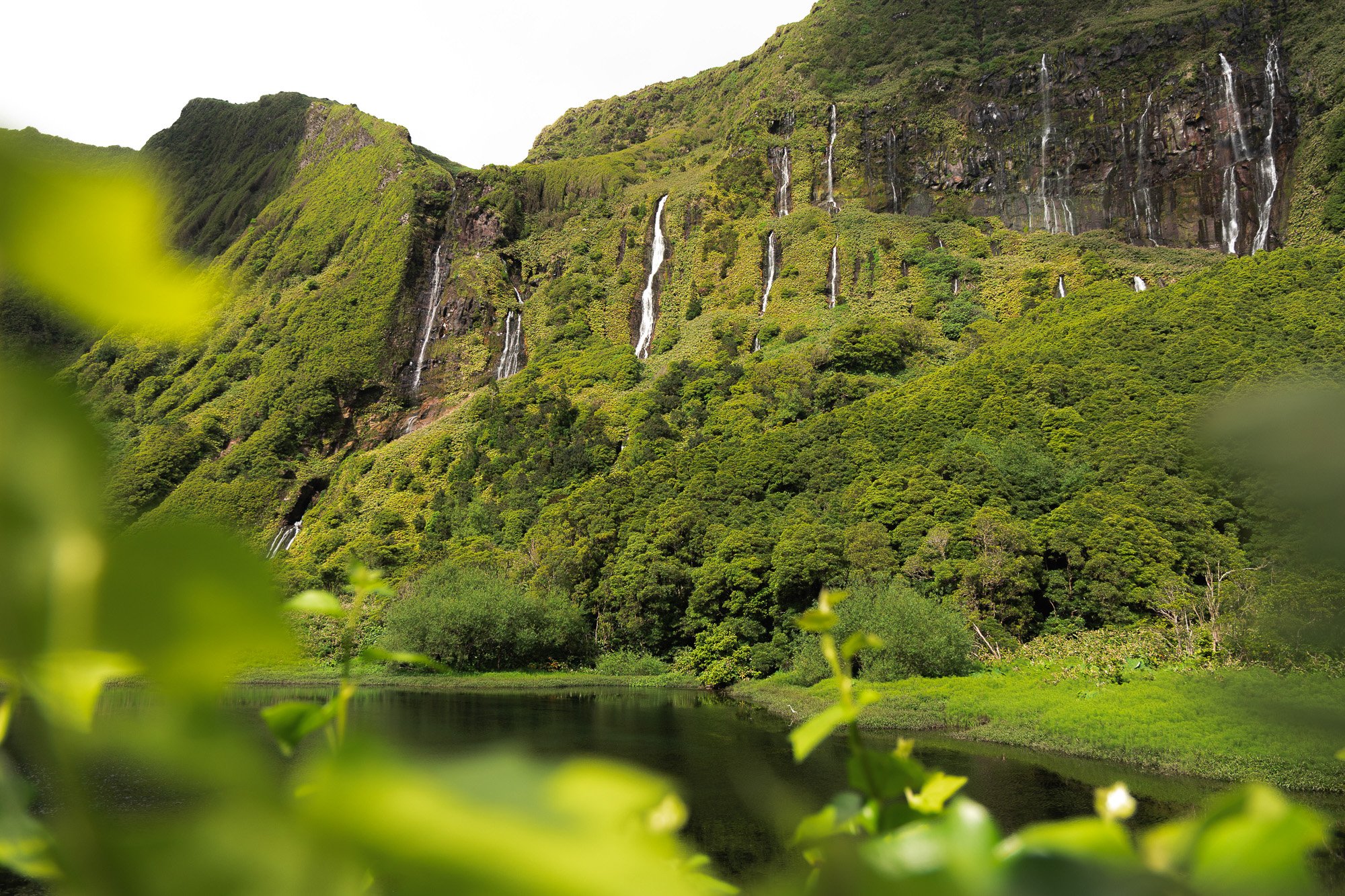 Grüne Berglandschaft mit mehreren Wasserfällen, ein See im Vordergrund, umgeben von Büschen und Bäumen.