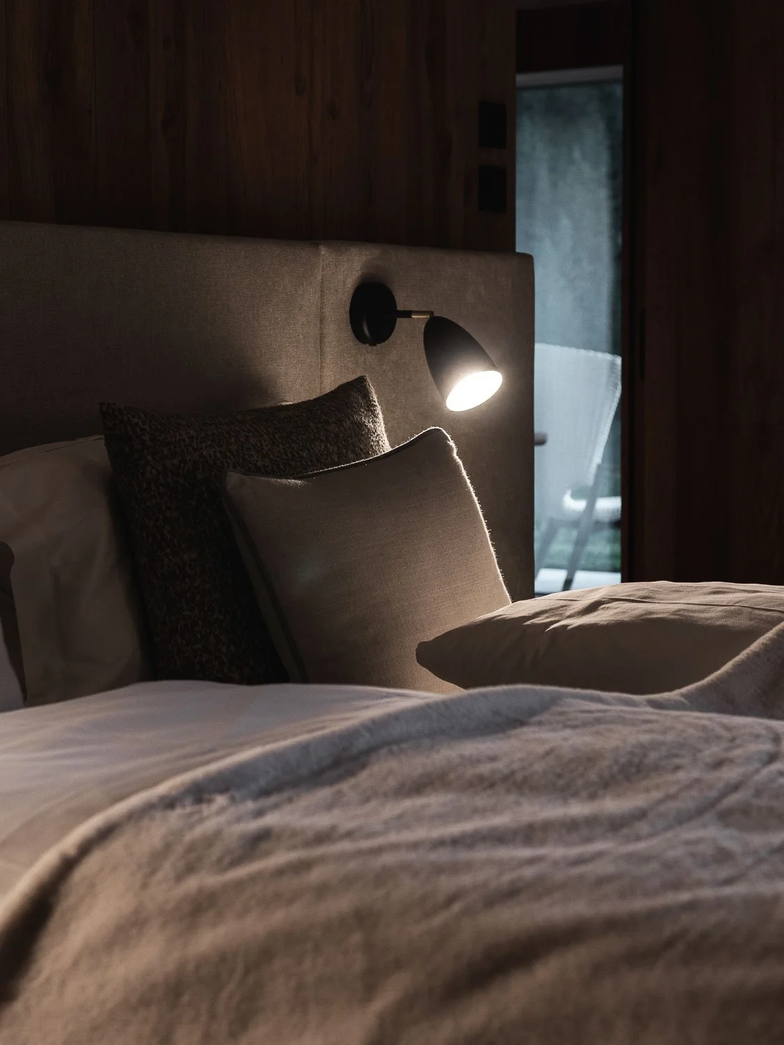 Ein Bett mit mehreren Kissen und einer Nachttischlampe in einem dunklen Schlafzimmer; im Hintergrund führt eine Glastür auf eine Terrasse.
