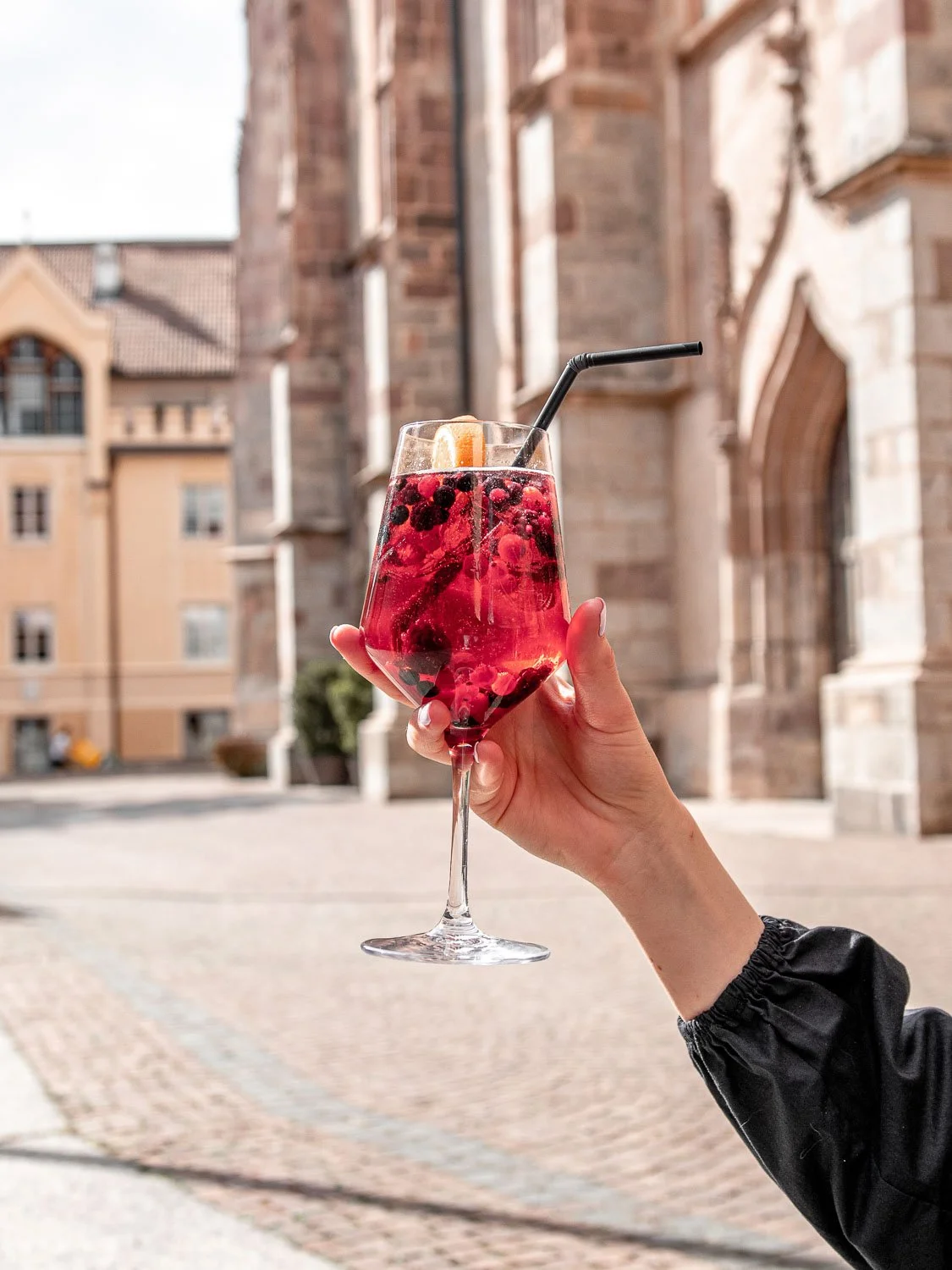 Person hält ein Glas mit roten Beeren und einer Zitronenscheibe vor einer historischen Steinarchitektur im Hintergrund.