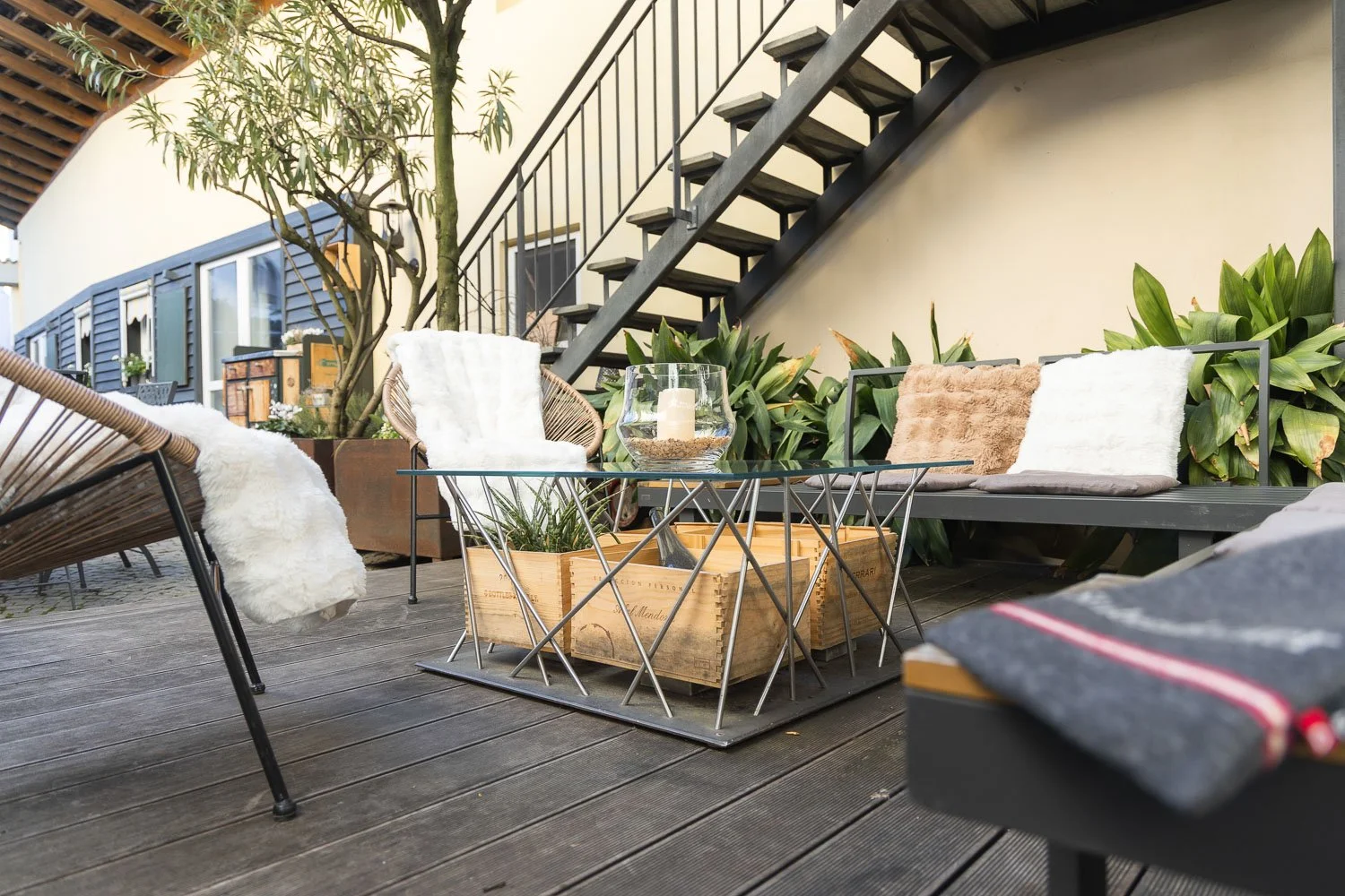 Gemütliche Terasse mit Holzmöbeln, Kissen, Pflanze, Kerze, und Stufen im Hintergrund, sonnige Atmosphäre.