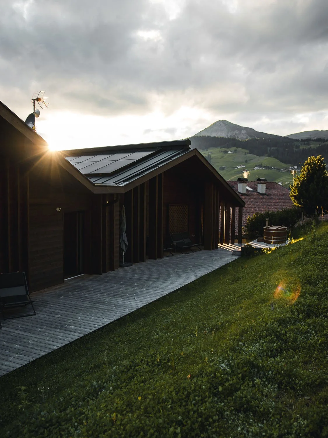 Ein Haus mit dunklem Holz und einer Terrasse, im Hintergrund Berge und ein bewölkter Himmel bei Sonnenuntergang.