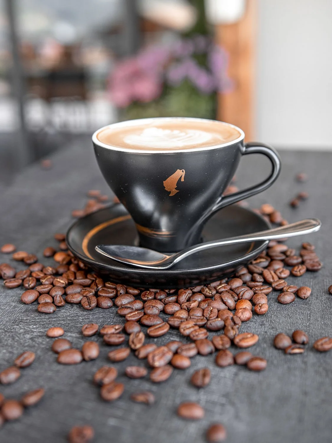 Schwarze Kaffeetasse mit Cappuccino und Milchschaum, umliegende Kaffeebohnen auf einem schwarzen Tisch, im Hintergrund blumen und ein Balkonfenster.