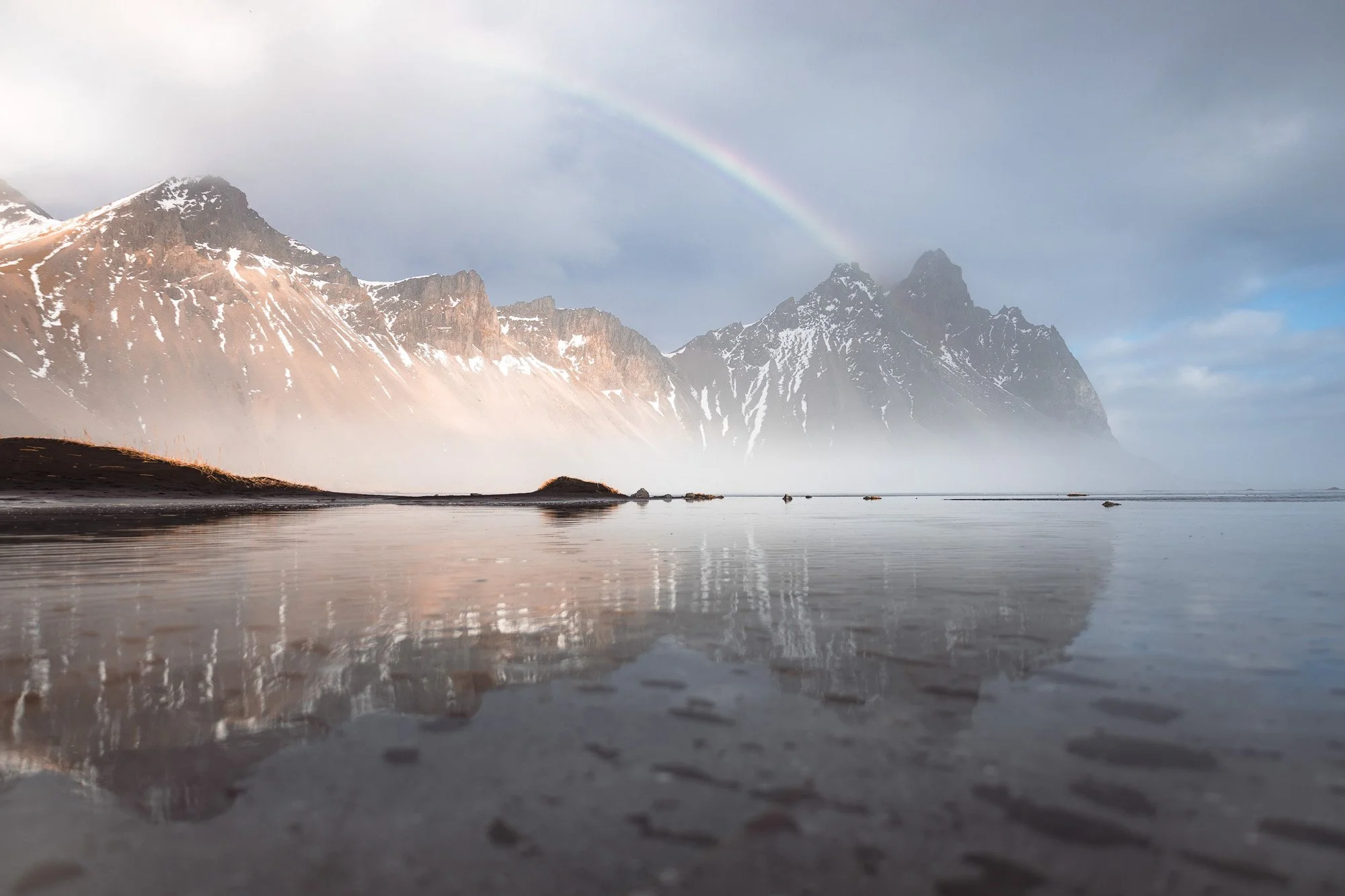 Berglandschaft mit verschneiten Gipfeln, Regenbogen, stürmischer Himmel und ruhiger See mit Reflexion der Berge.