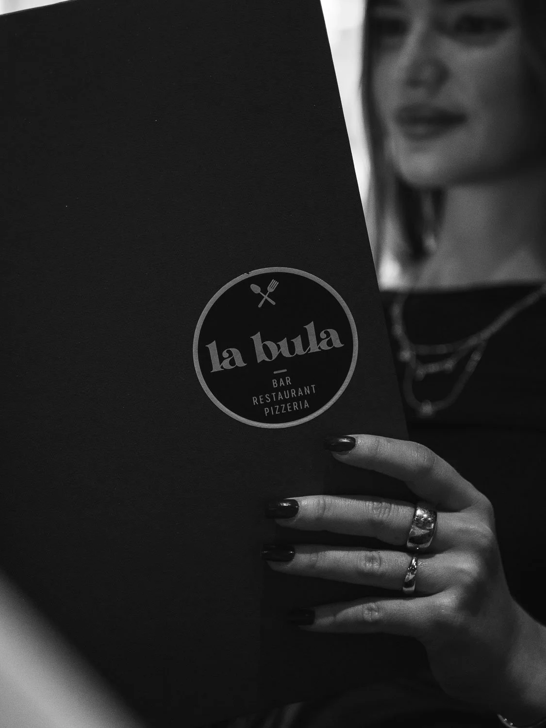 Nahaufnahme eines schwarzen Menüs mit dem Logo "la bula" für Bar, Restaurant und Pizzeria, gehalten von einer Person mit schwarzen Nägeln und mehreren Ringen, im Schwarz-Weiß-Foto.