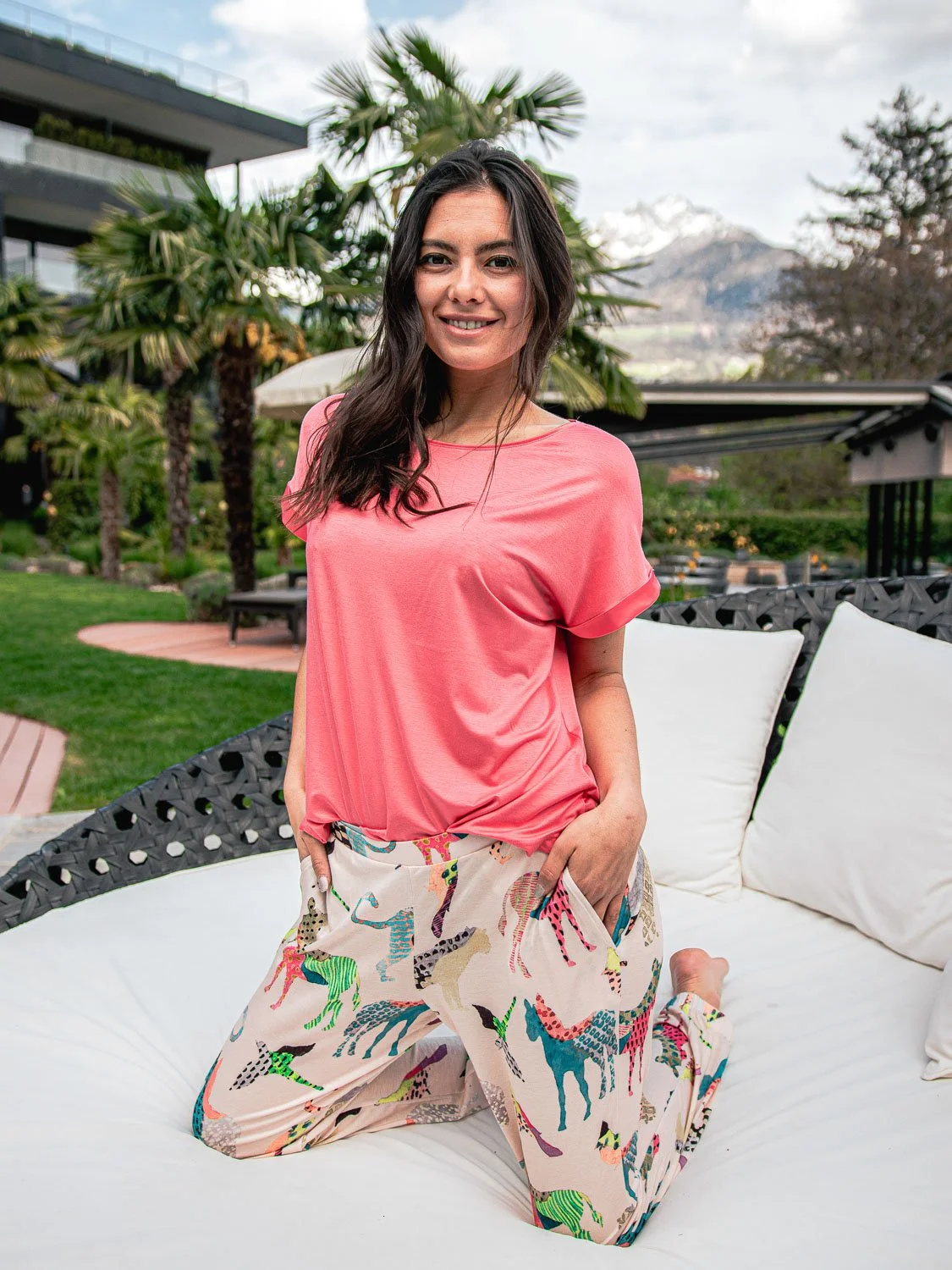 Frau in pinkem Top und bunten Haremshosen, knieend auf einem weißen Sitzkissen im Garten.