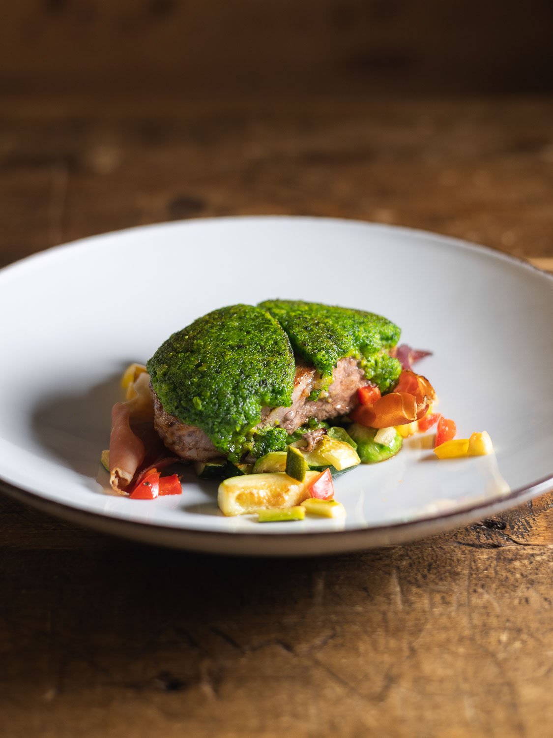 Ein Teller mit einem Fleischpatty, bedeckt mit grünem Brokkoli, serviert mit klein geschnittenem Gemüse auf einem weißen Teller auf einem Holztablett.