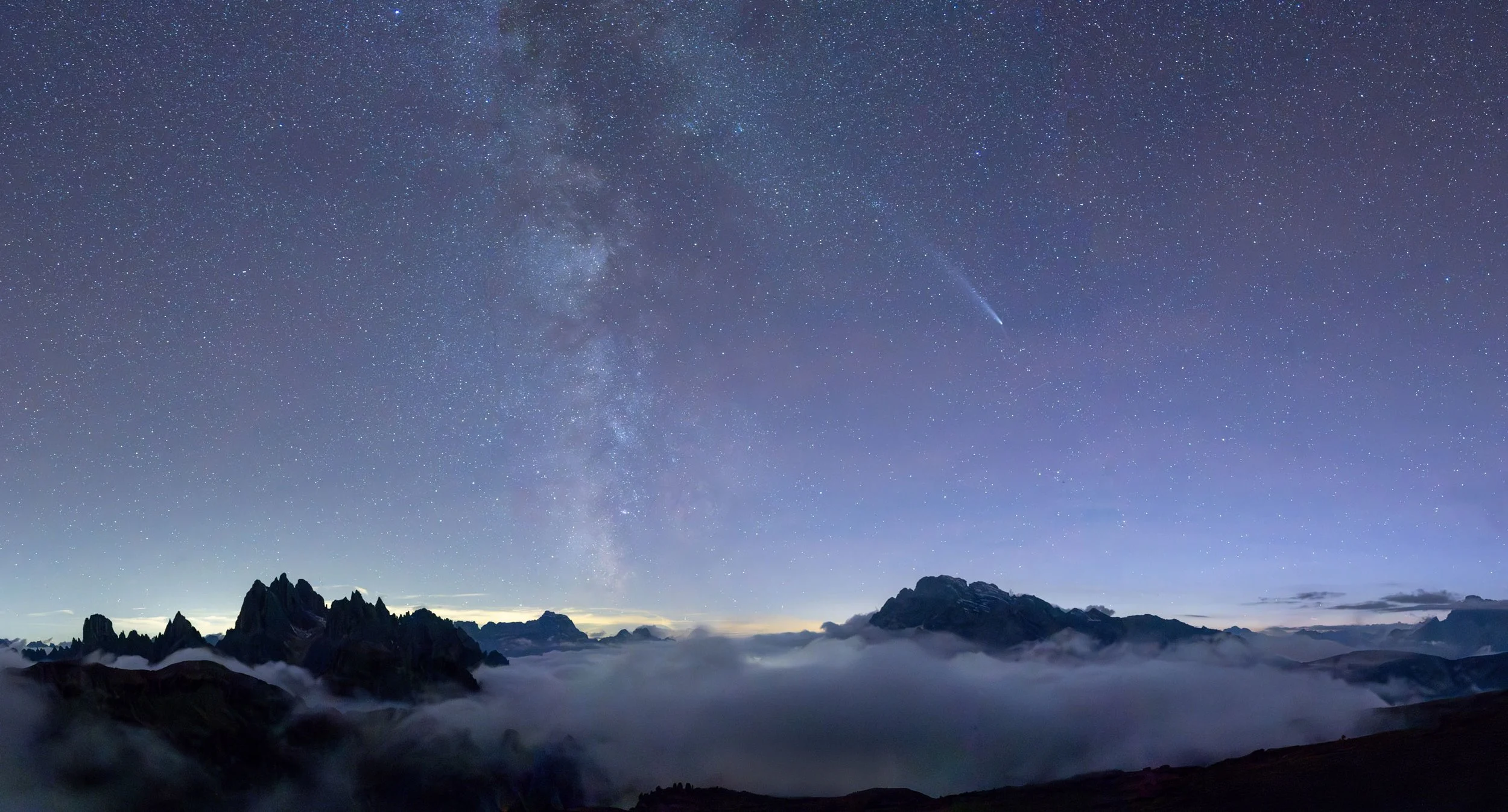 Sternenhimmel mit Milchstraße und Komet über verschneiten Bergen bei Nacht, mit Wolken im Tal.
