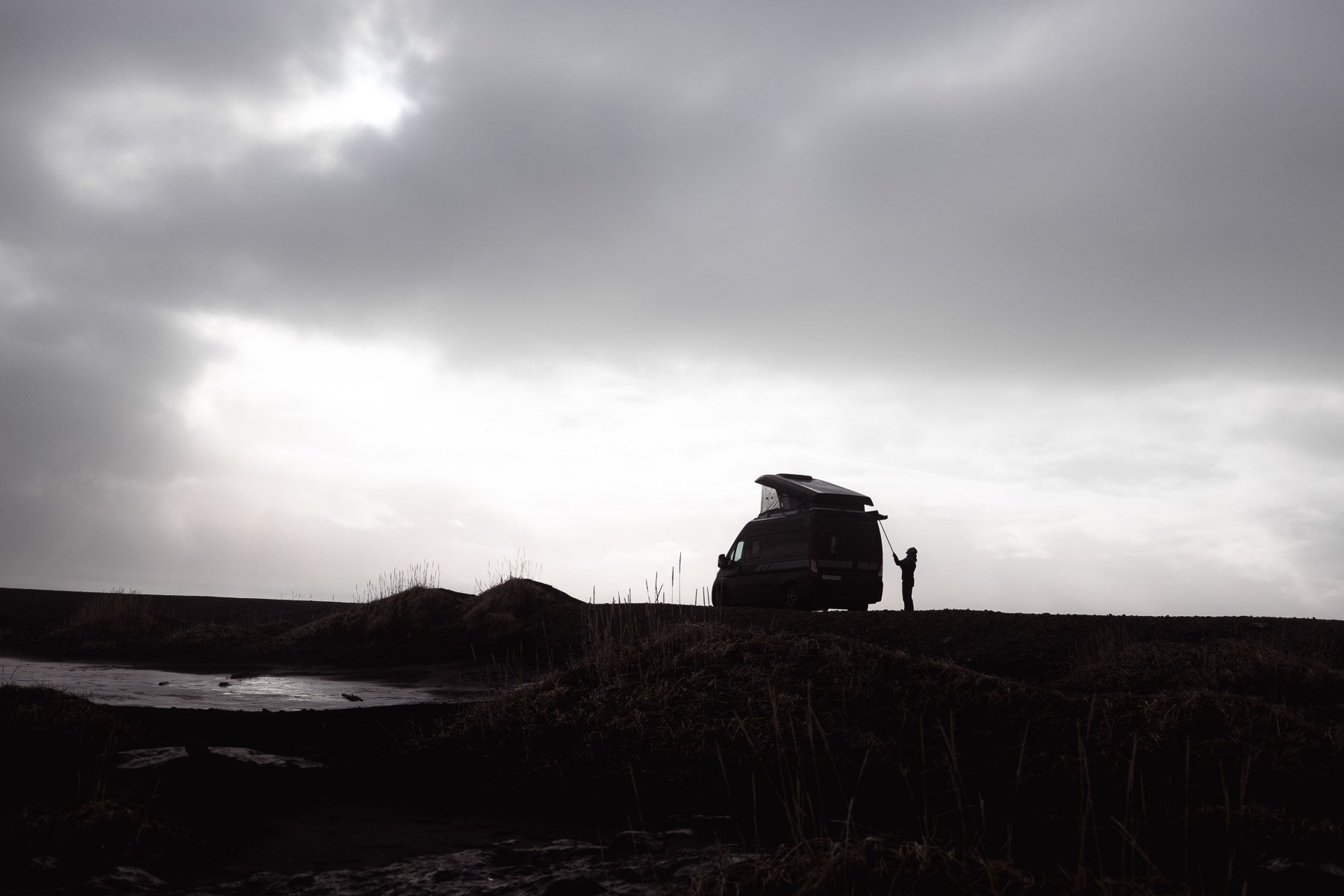 Silhouette eines Menschen steht neben einem Campervan auf einer dunklen, unberührten Landschaft bei bewölktem Himmel.