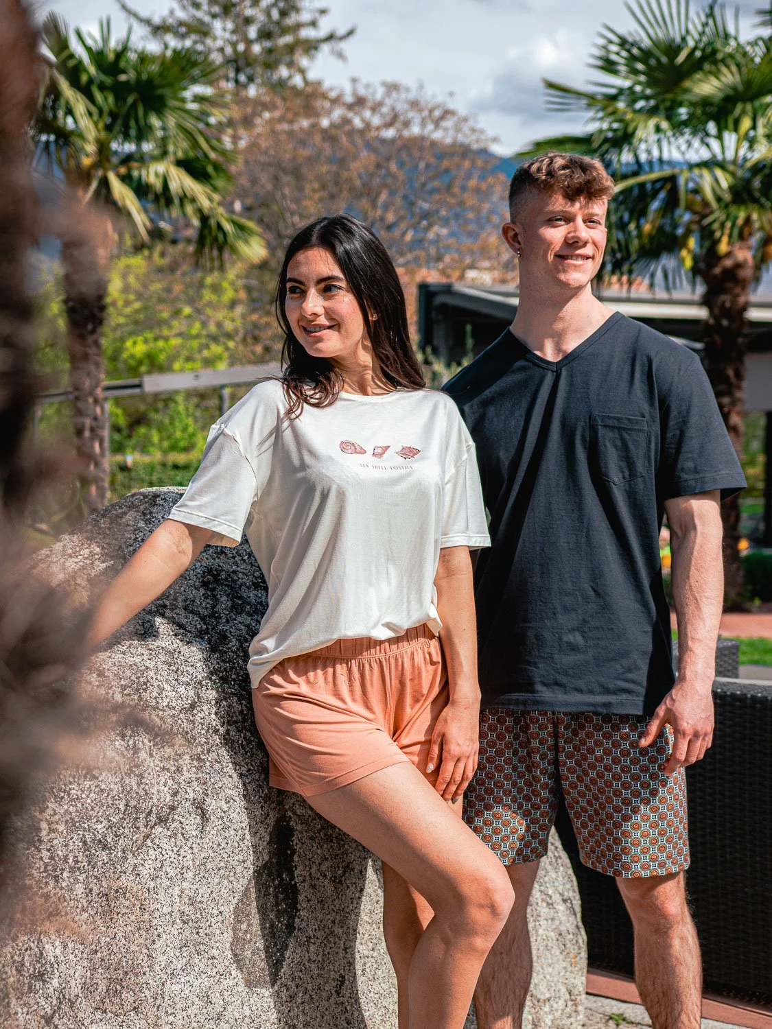 Ein Mann und eine Frau stehen im Freien vor Bäumen und einem großen Felsen, lächelnd und entspannt.