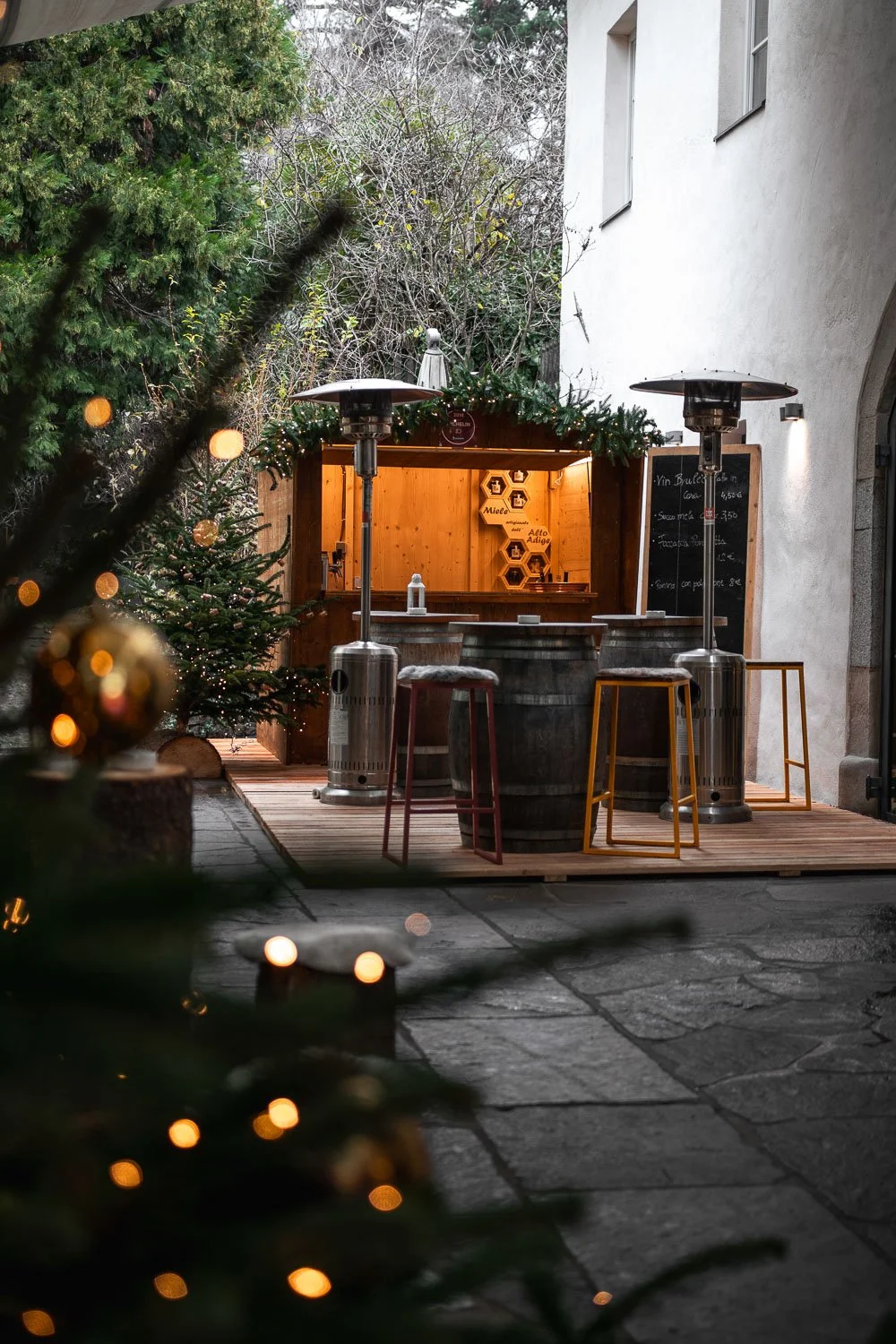 Ein festlich dekorierter Holzstand mit Stehtischen und Heizstrahlern, im Hintergrund Tannenbäume und eine dunkle, steinige Hoffläche.