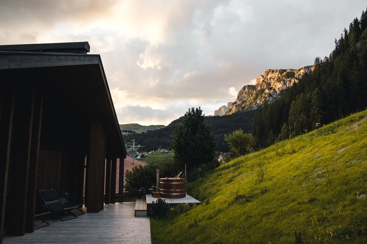 Blick auf eine Hügellandschaft mit einem Holzhaus im Vordergrund, einem Baum, einem Holzbadewanne und einer grünen, bewaldeten Bergkette im Hintergrund bei bewölktem Himmel.