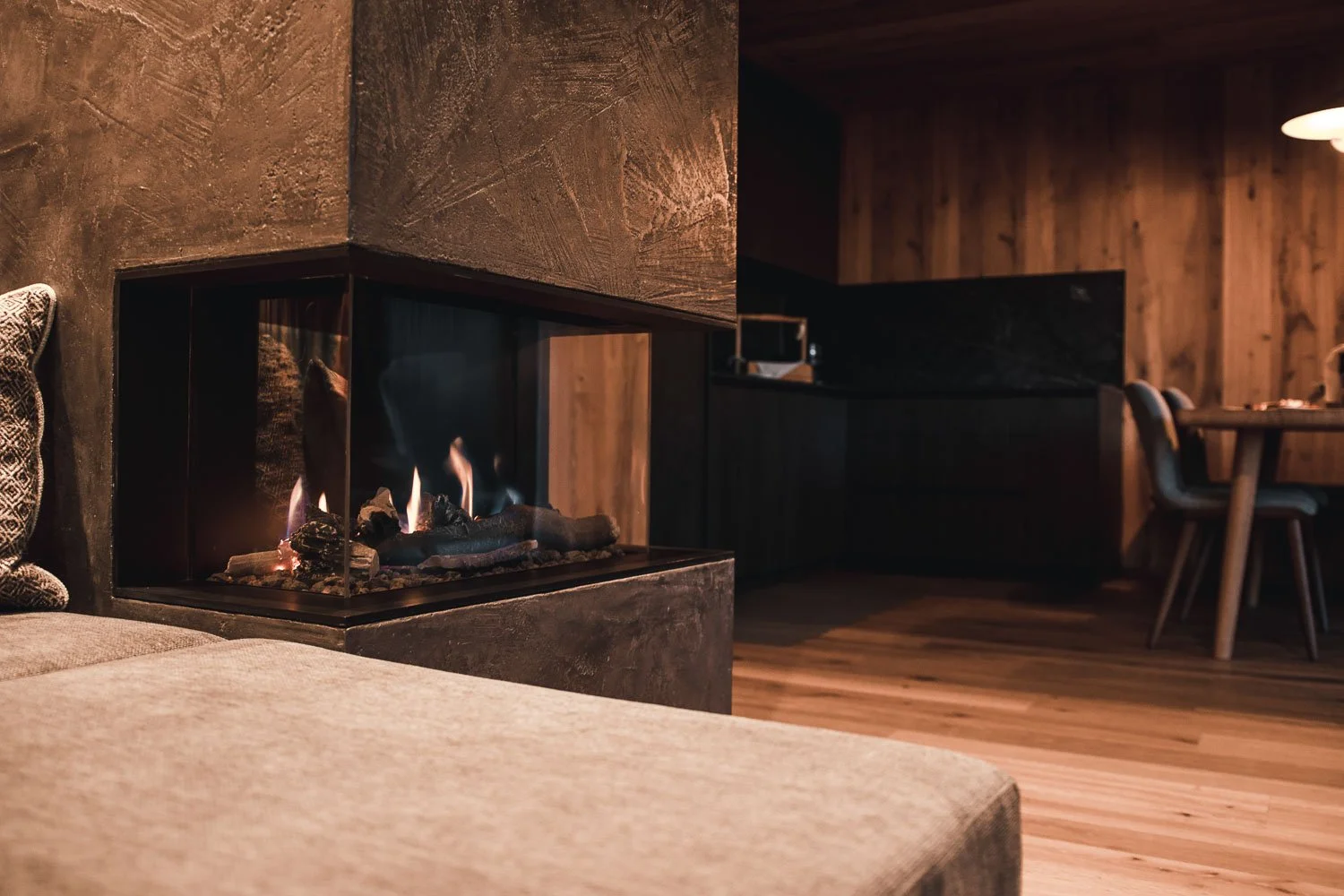 Moderner Wohnraum mit sichtbarem Kamin, braune Holzwände, Holzfußboden, beige Couch und dunkle Einrichtungsgegenstände.