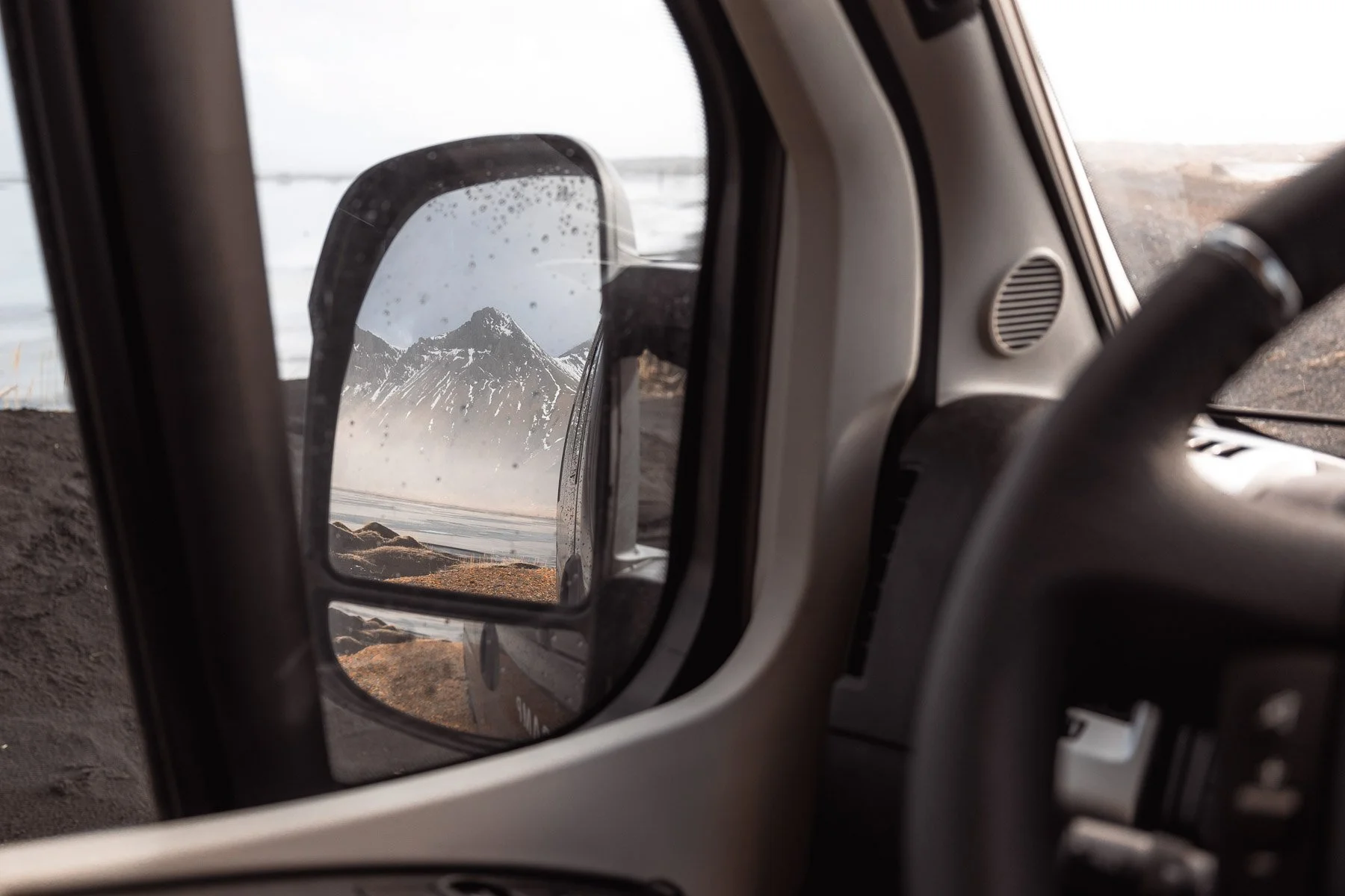 Berg mit schneebedecktem Gipfel, im Hintergrund Wasser und ein wolkenbedeckter Himmel, aufgenommen durch den Seitenspiegel eines Vans in einer kalten, rauen Umgebung.