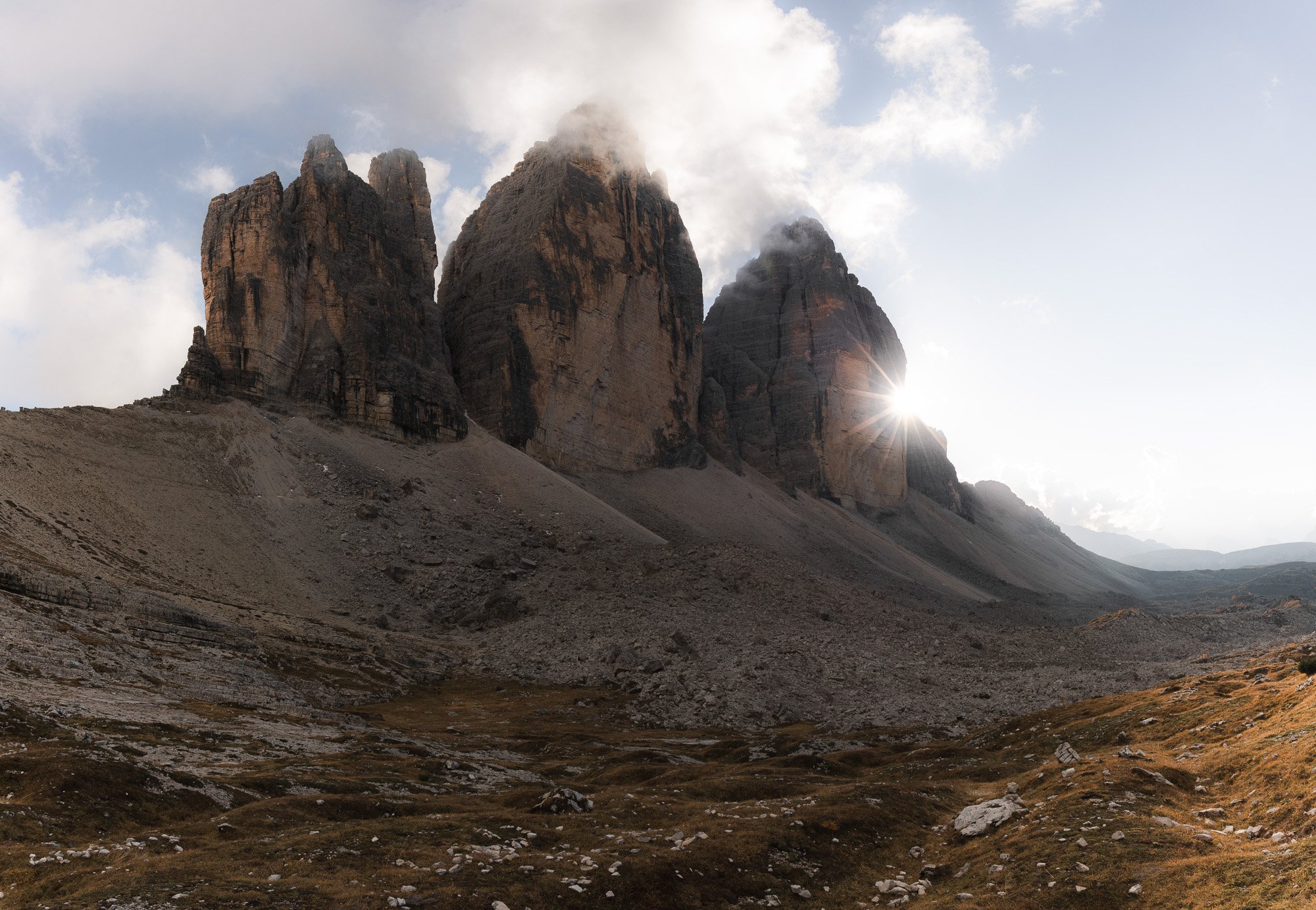 Drei große, felsige Berge mit Wolken am Himmel, Sonne hinter dem rechten Berg, umgeben von einer kargen, hügeligen Landschaft.