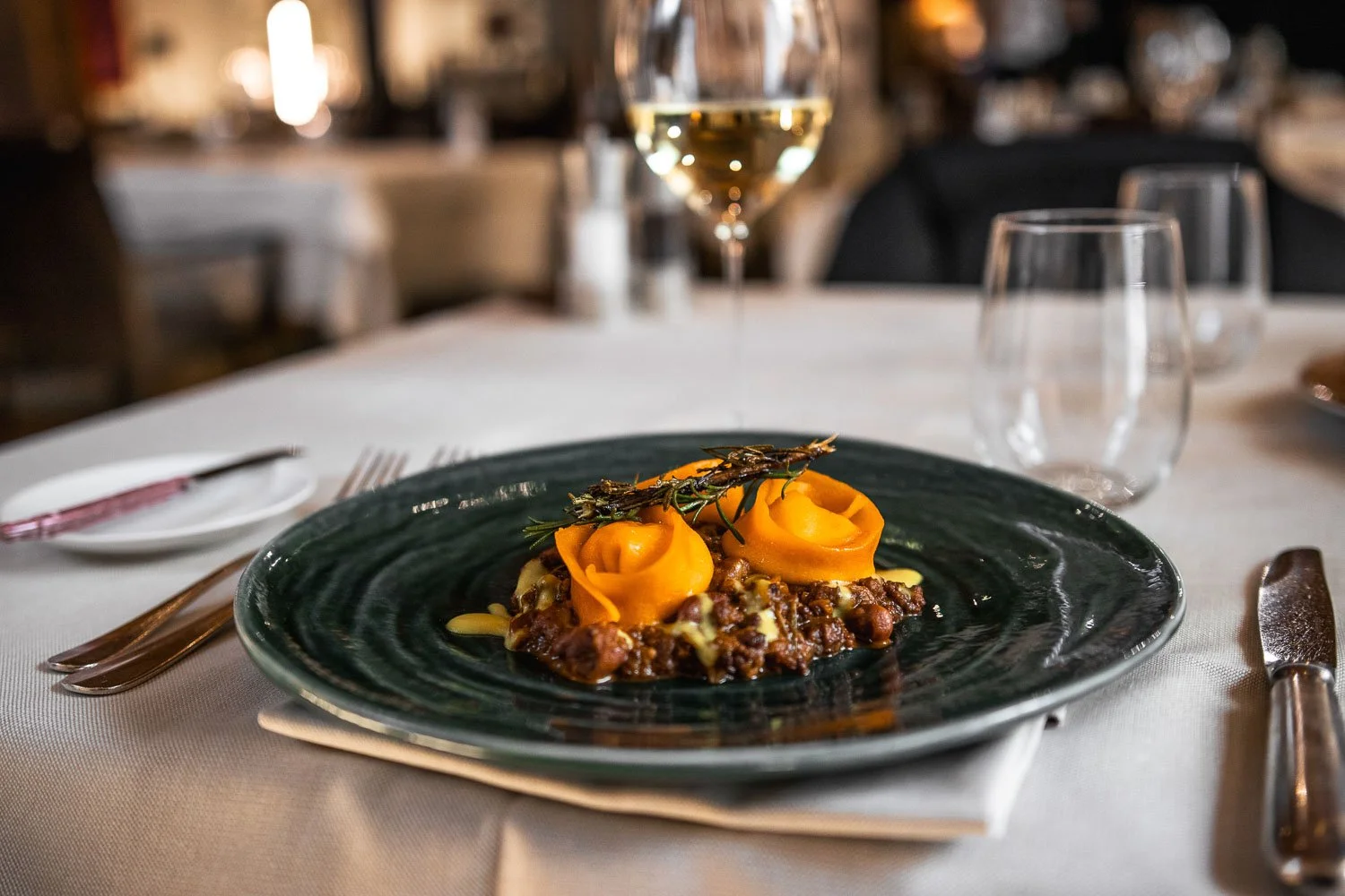 Hauptgang auf einem weißen Tisch, dunkler Teller mit zwei gefüllten, gelben, pastellfarbenen Teigtaschen, garnieren mit einer kleinen Zweig Rosmarin und Sommerlapen, im Hintergrund ein Glas Weißwein und weiteres Tischgeschirr.