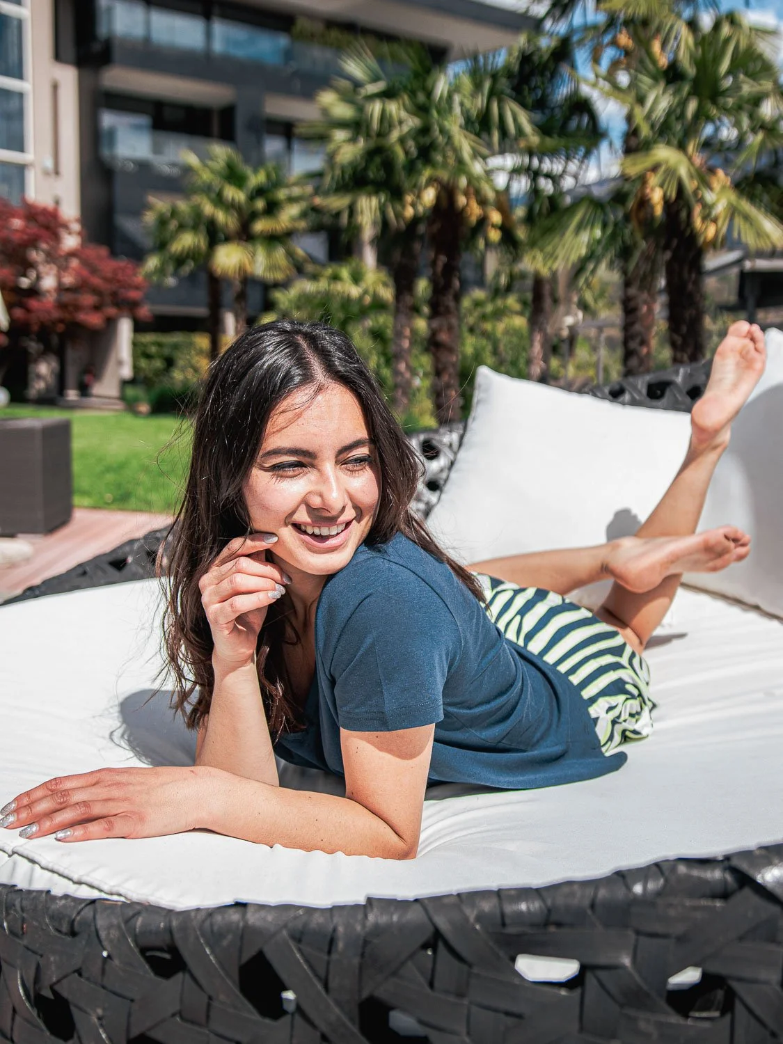 Junge Frau liegt lachend auf einem großen weißen Kissen im Freien, umgeben von Palmen, an einem sonnigen Tag.