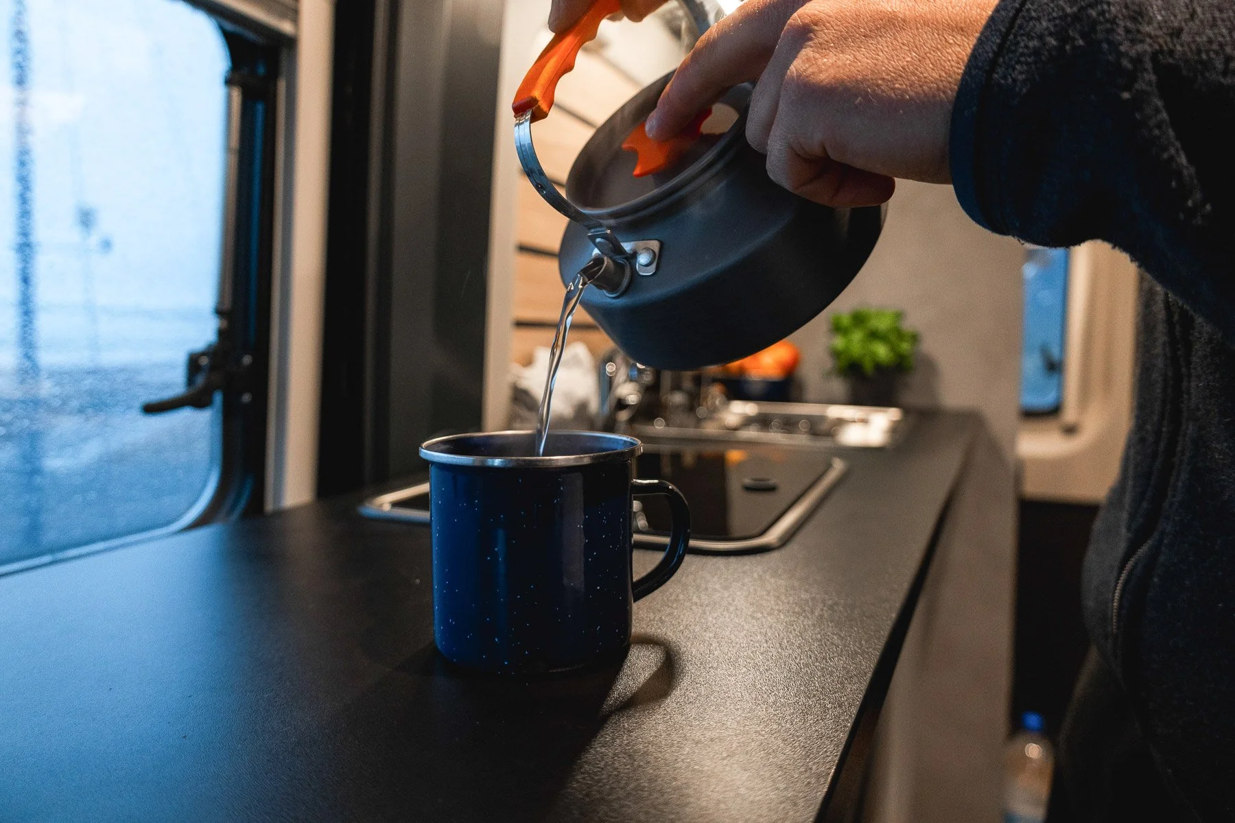 Eine Person gießt Wasser aus einem schwarzen Wasserkocher in eine dunkle Tasse auf einer Küchenarbeitsplatte im Wohnmobil.