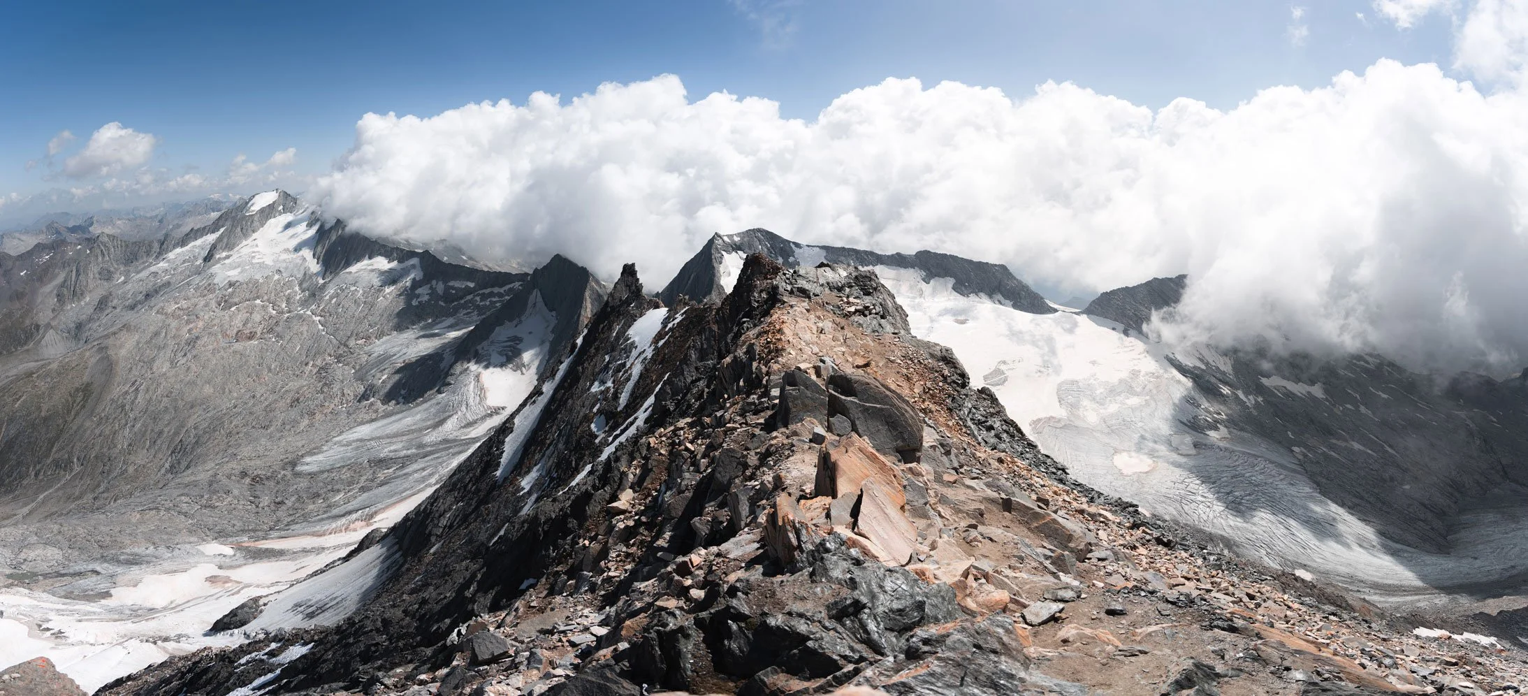 Hochalpine Berggipfel mit Felsen und Eis, Wolken am Himmel, in der Ferne weitere Berge.