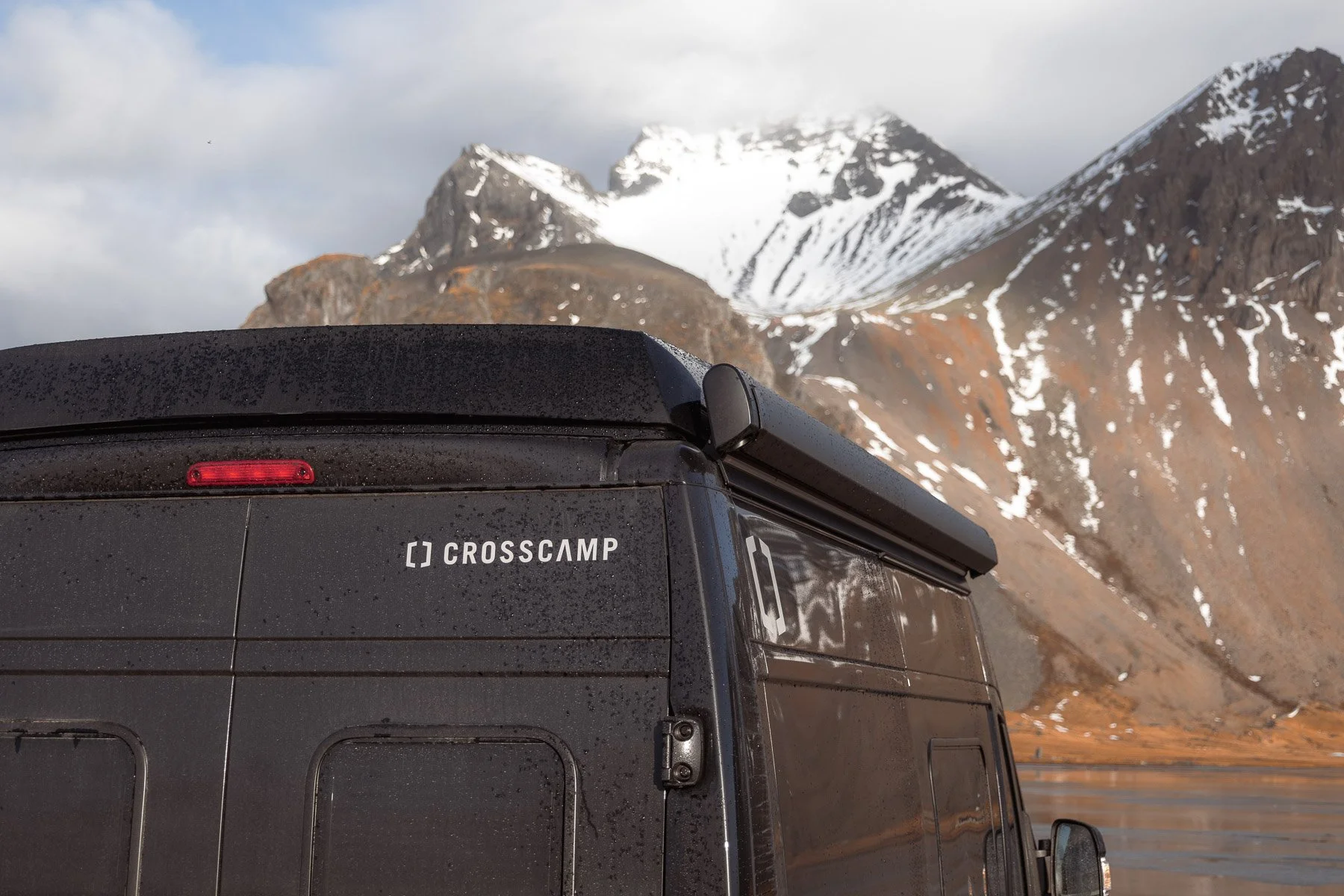 Nahaufnahme von einem schwarzen Van mit der Aufschrift 'Crosscamp' im Vordergrund, mit Wassertröpfchen auf der Oberfläche und einer Berglandschaft mit verschneiten Gipfeln im Hintergrund.