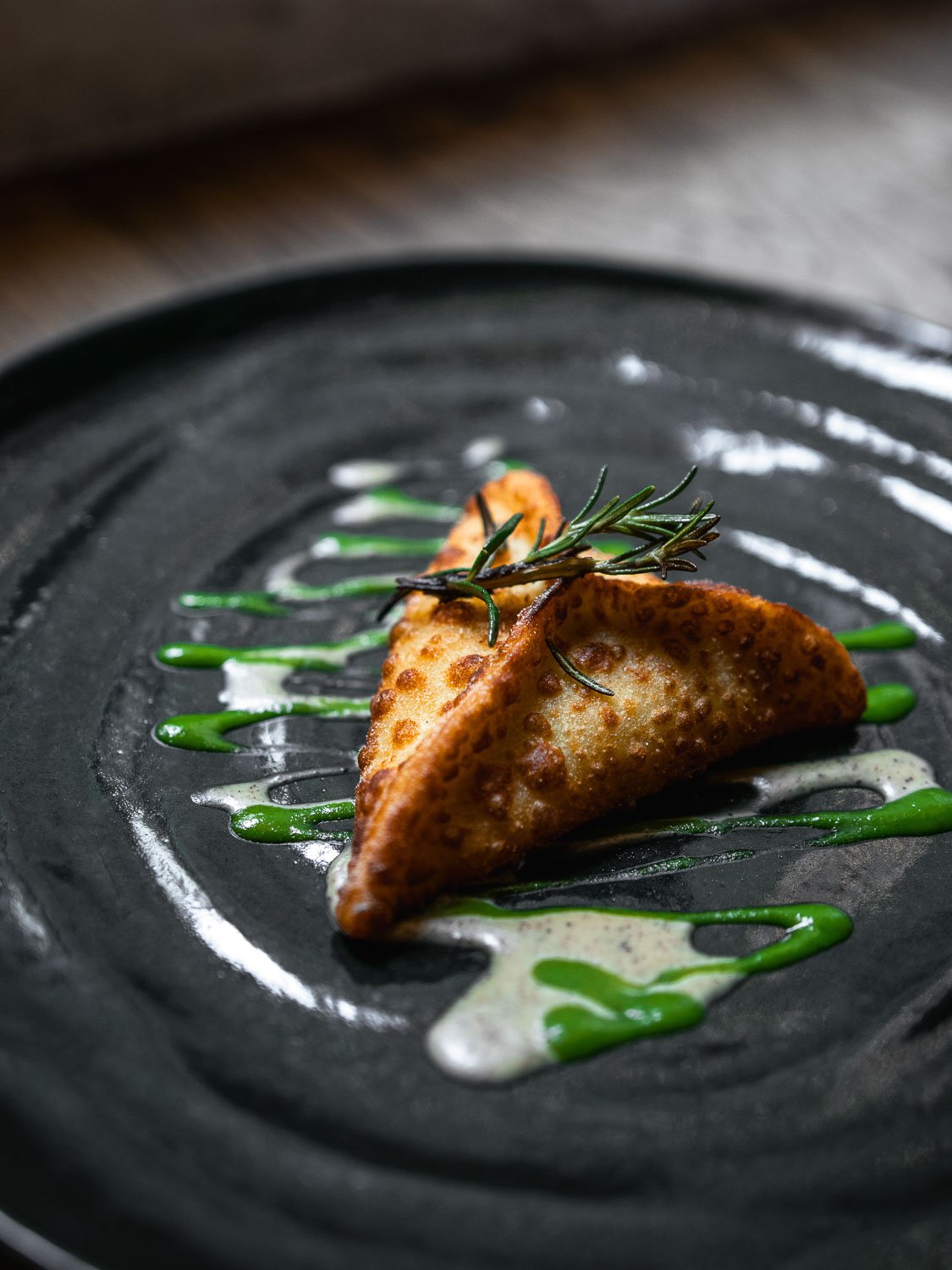 Ein gebratener Teigtasche mit Kräutern auf einem schwarzen Teller, garniert mit grüner Sauce und weißer Sauce.