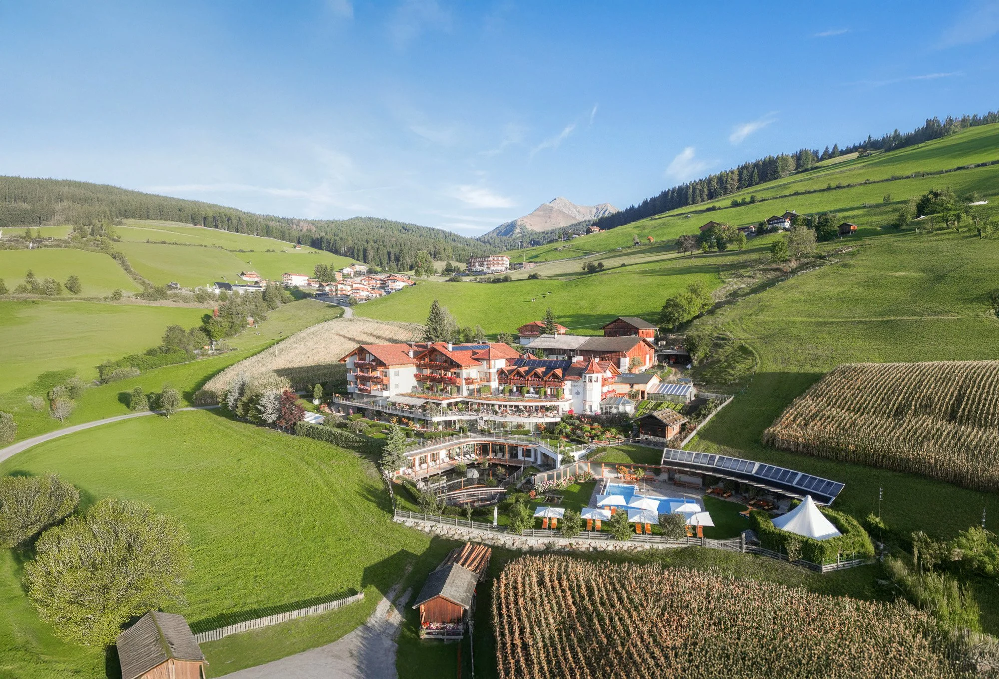 Landschaftliche Drohnenaufnahme mit grünen Hügeln, einem Dorf mit rot-weißen Gebäuden und einem Hotel mit Pool, umgeben von landwirtschaftlichen Flächen und Bäumen unter einem blauen Himmel.