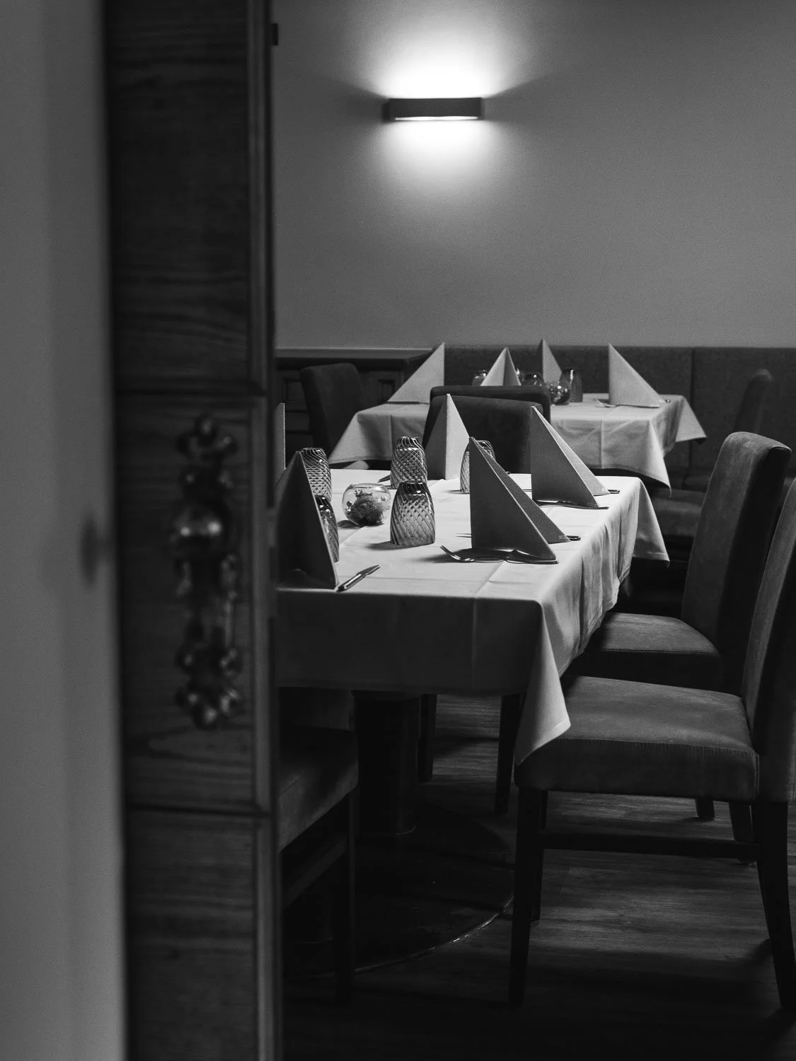 Ein eleganter, gedeckter Esstisch mit weißen Tischtüchern, Stoffservietten, Kerzen und Wassergläsern in einem dunklen, gedämpften Restaurant. Das Bild ist in Schwarz-Weiß gehalten.