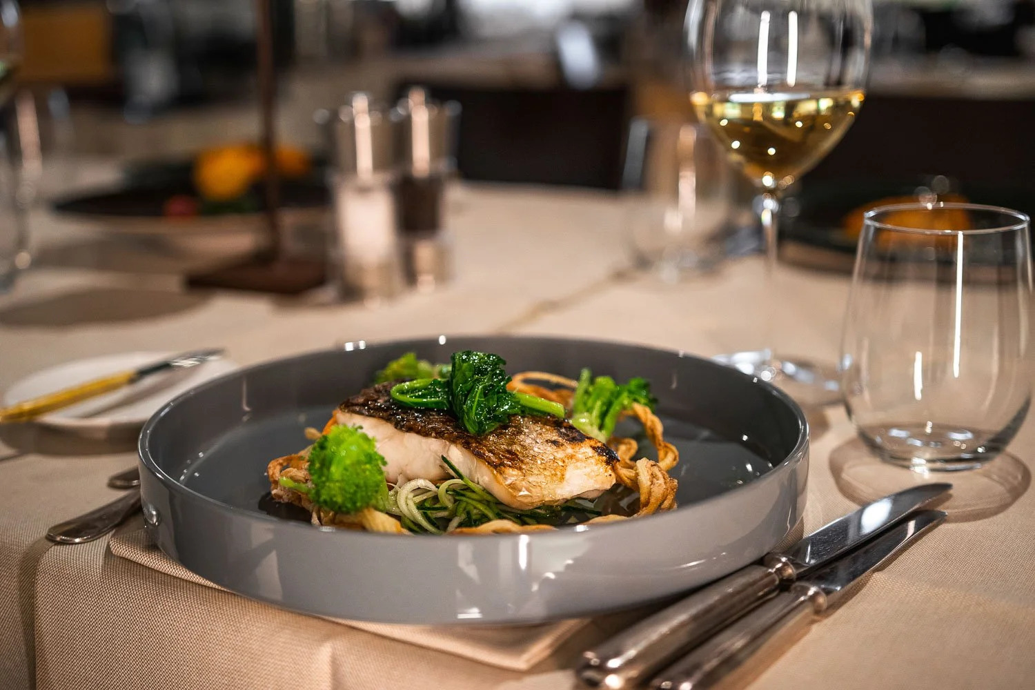 Fischgericht mit Gemüse auf einem schwarzen Teller, auf einem beige gedeckten Tisch, mit Weinglas, Wasser- und Weinglas, Besteck und Salz- und Pfeffermühlen im Hintergrund.