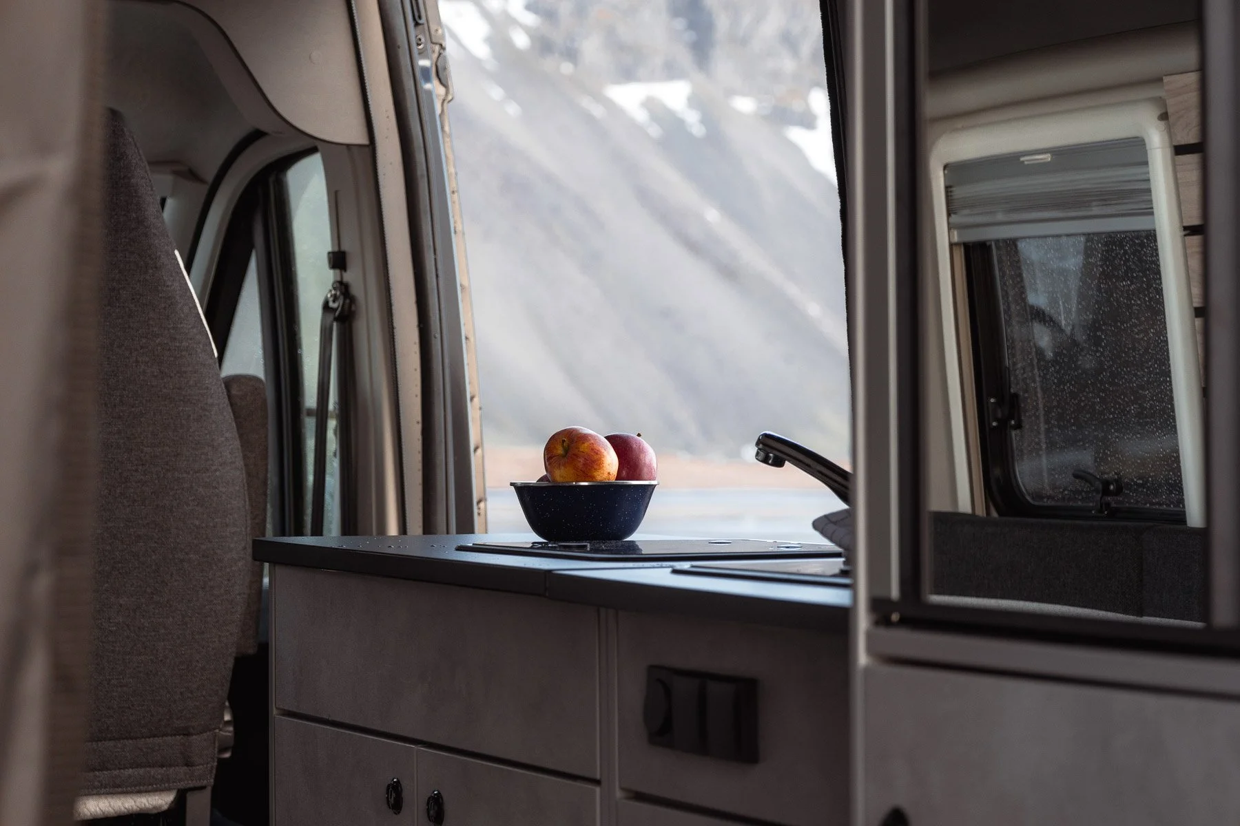 Innenansicht eines Wohnmobils mit einem Blick auf die Küchenzeile und ein Fenster, auf dem eine Schüssel mit Äpfeln steht, im Hintergrund eine bergige Landschaft.