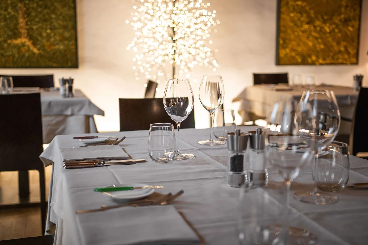Ein gedeckter Esstisch in einem eleganten Restaurant mit Weißem Tischtuch, Weingläsern, Wasserbechern, Besteck, Salz- und Pfefferstreuern, in einem Raum mit dunklen Stühlen, moderner Kunst an der Wand, und einer dekorativen beleuchteten Baumskulptur 
