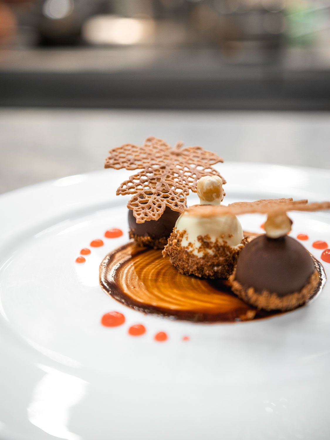 Dekoratives Dessert mit zwei Schokoladenkugeln, einer weißen Schokoladenkugel, einer essbaren Dekoration, einem essbaren Blatt, roten Soßenpunkten und einem kunstvollen Schokoladenmuster auf einem weißen Teller.