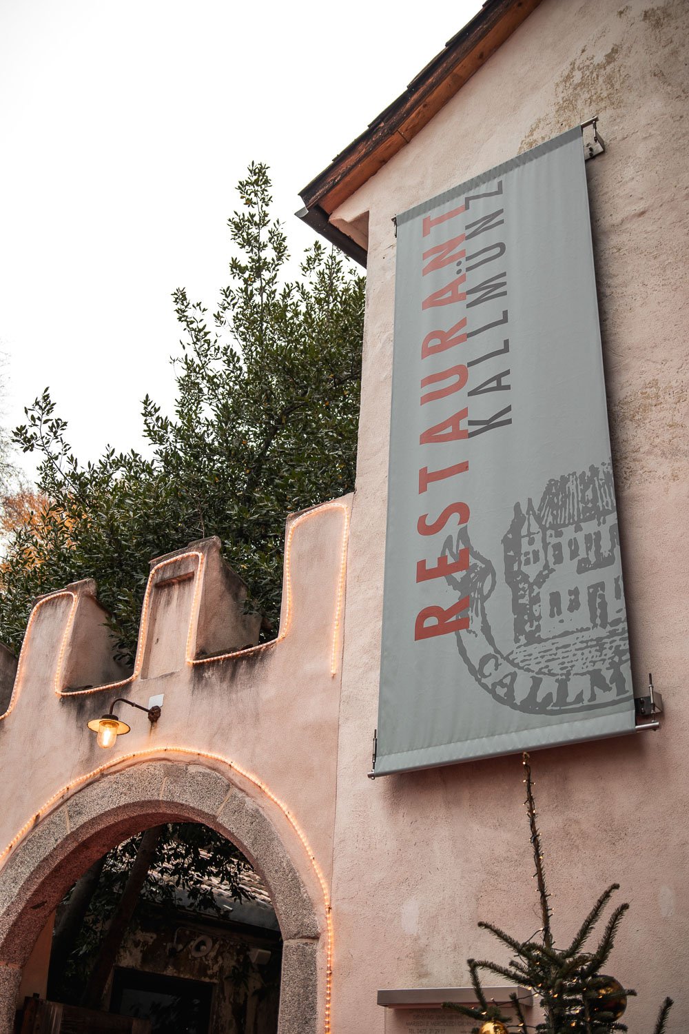 Ein vertikaler Banner mit der Aufschrift 'RESTAURANT KALLMÜNZ' an einer hellen Hauswand, mit einem Tor und Weihnachtsdekoration im Vordergrund, und Bäumen im Hintergrund.